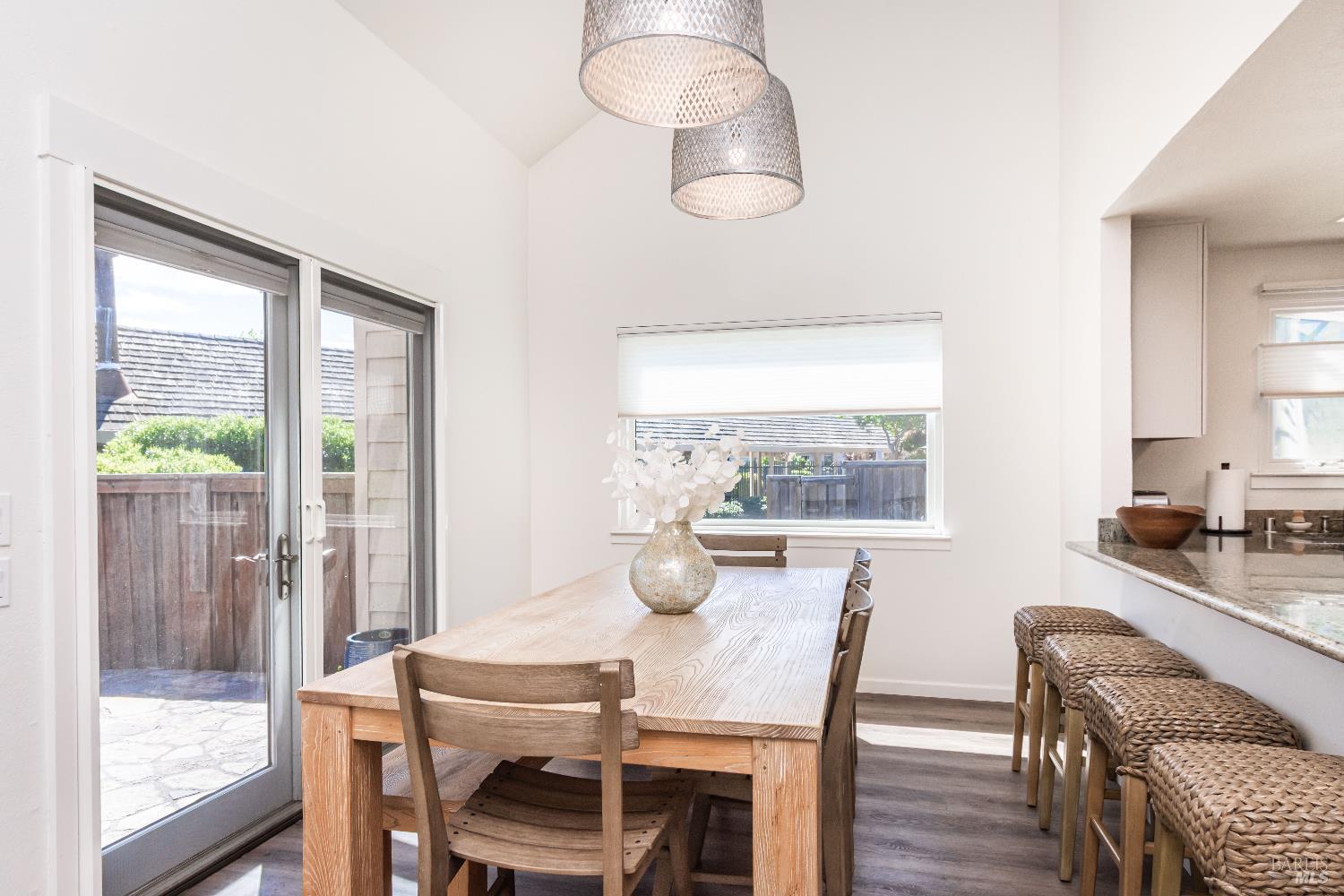 Dining area with a long wooden table, woven pendant lights, and kitchen counter stools beside sliding glass doors.