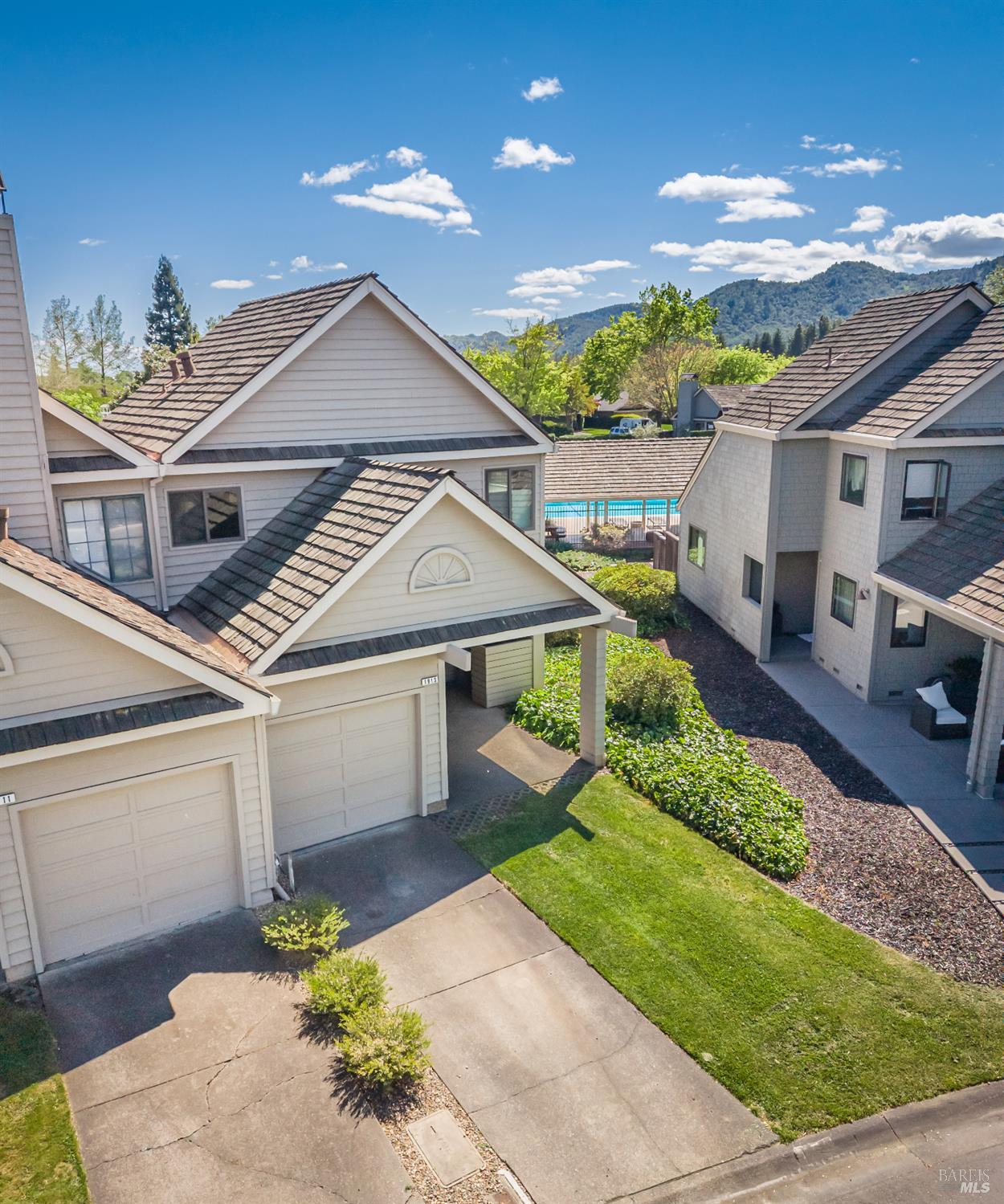 Neighboring homes with tiled roofs, a swimming pool, landscaped lawns, and wooded mountains beyond.