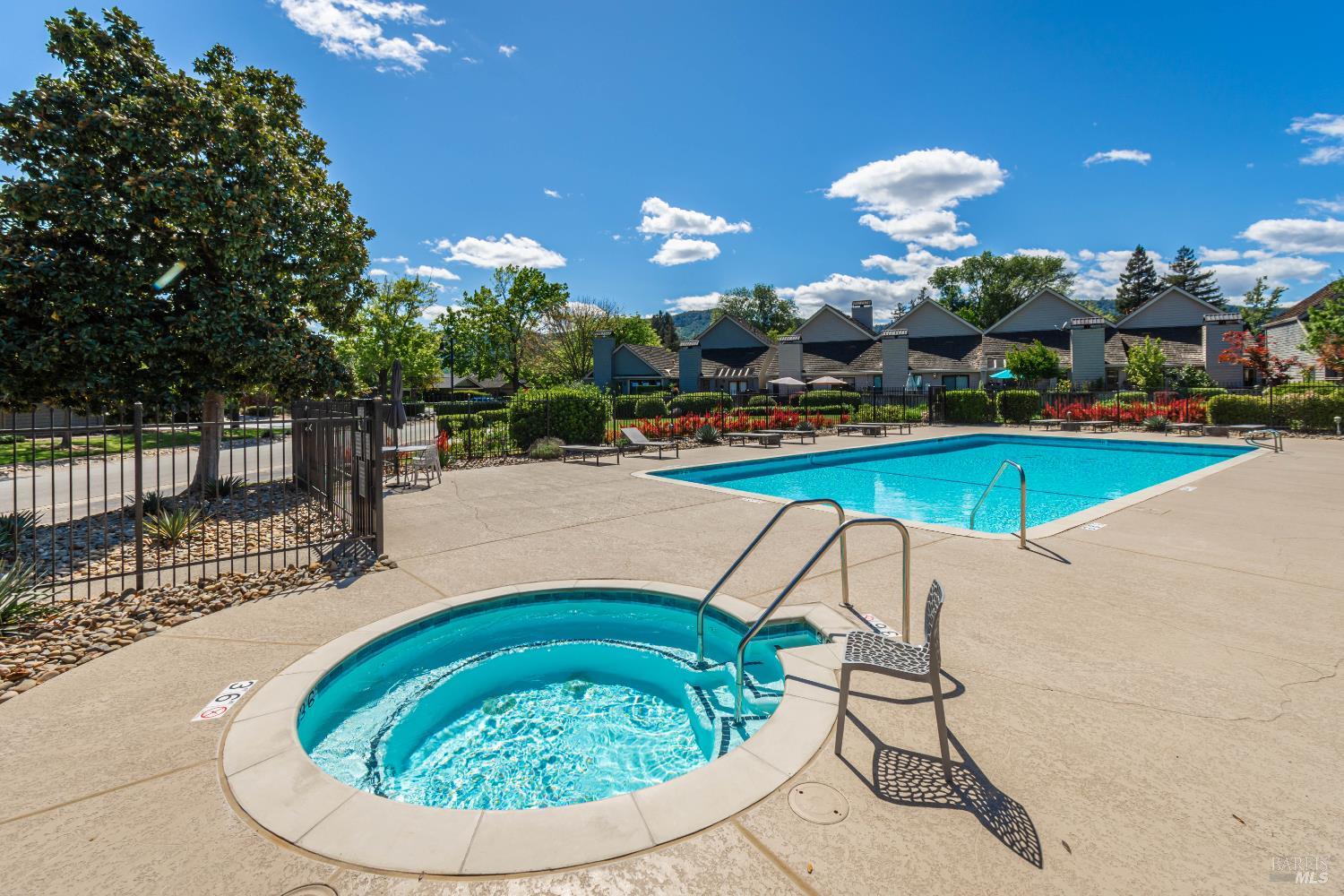 Community pool and circular spa surrounded by a paved deck, lounge chairs, flowers, and homes.