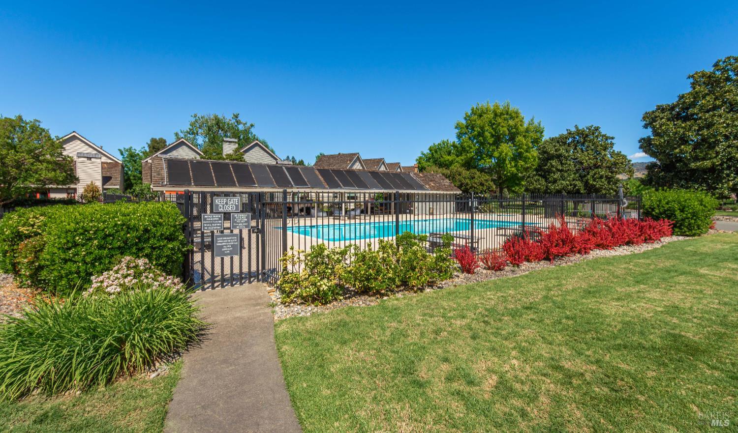 Fenced community swimming pool with a turquoise pool, landscaped lawn, and residential buildings behind it.
