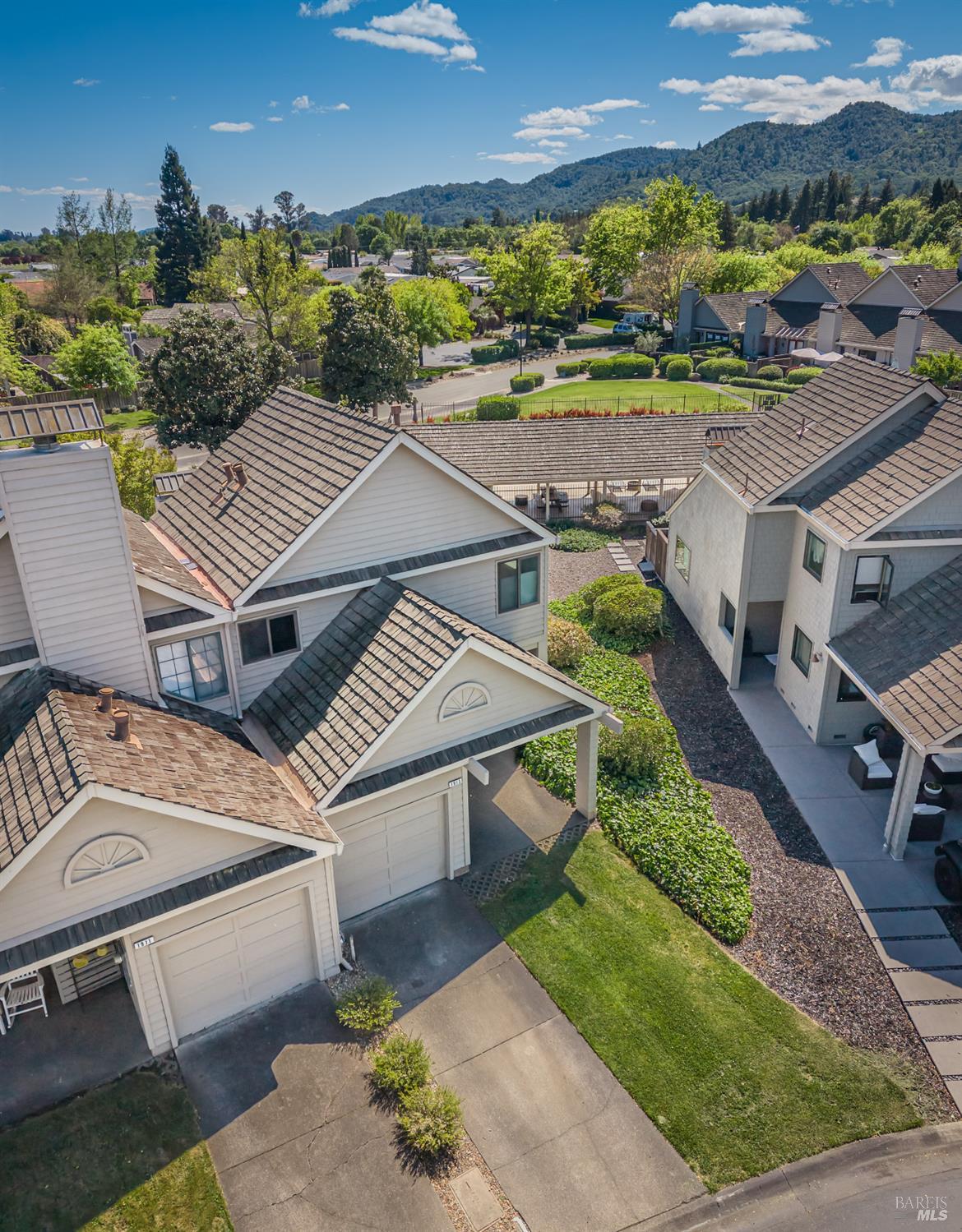 Aerial view of a home with neighboring houses, landscaped lawns, and forested hills beyond.