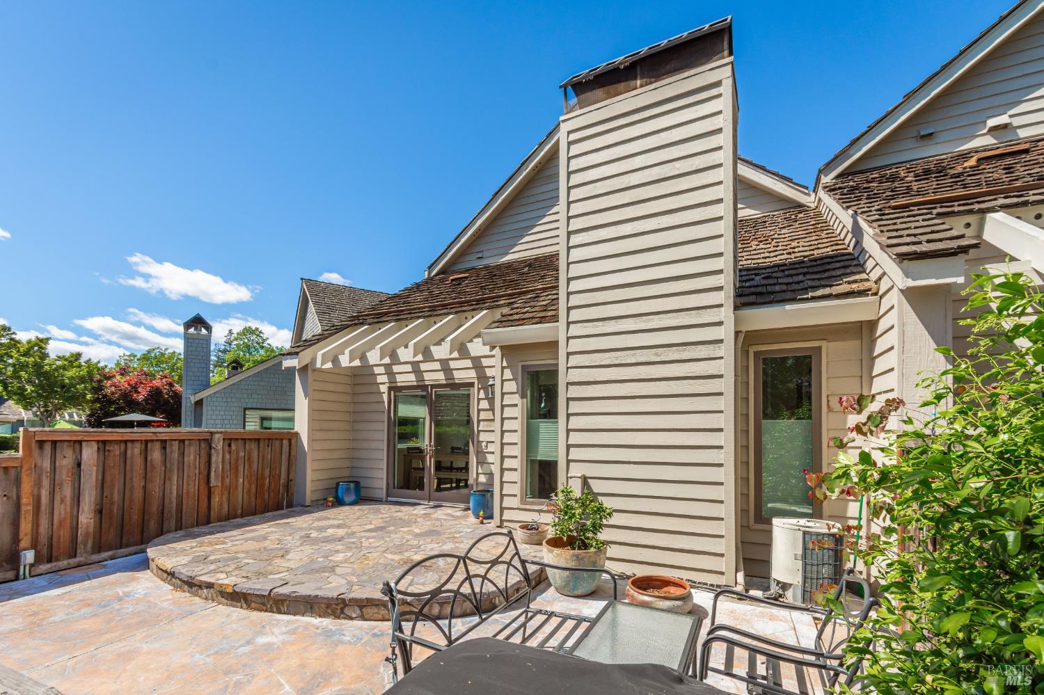 House exterior with a stone patio, black metal seating, potted plants, and a wooden fence.