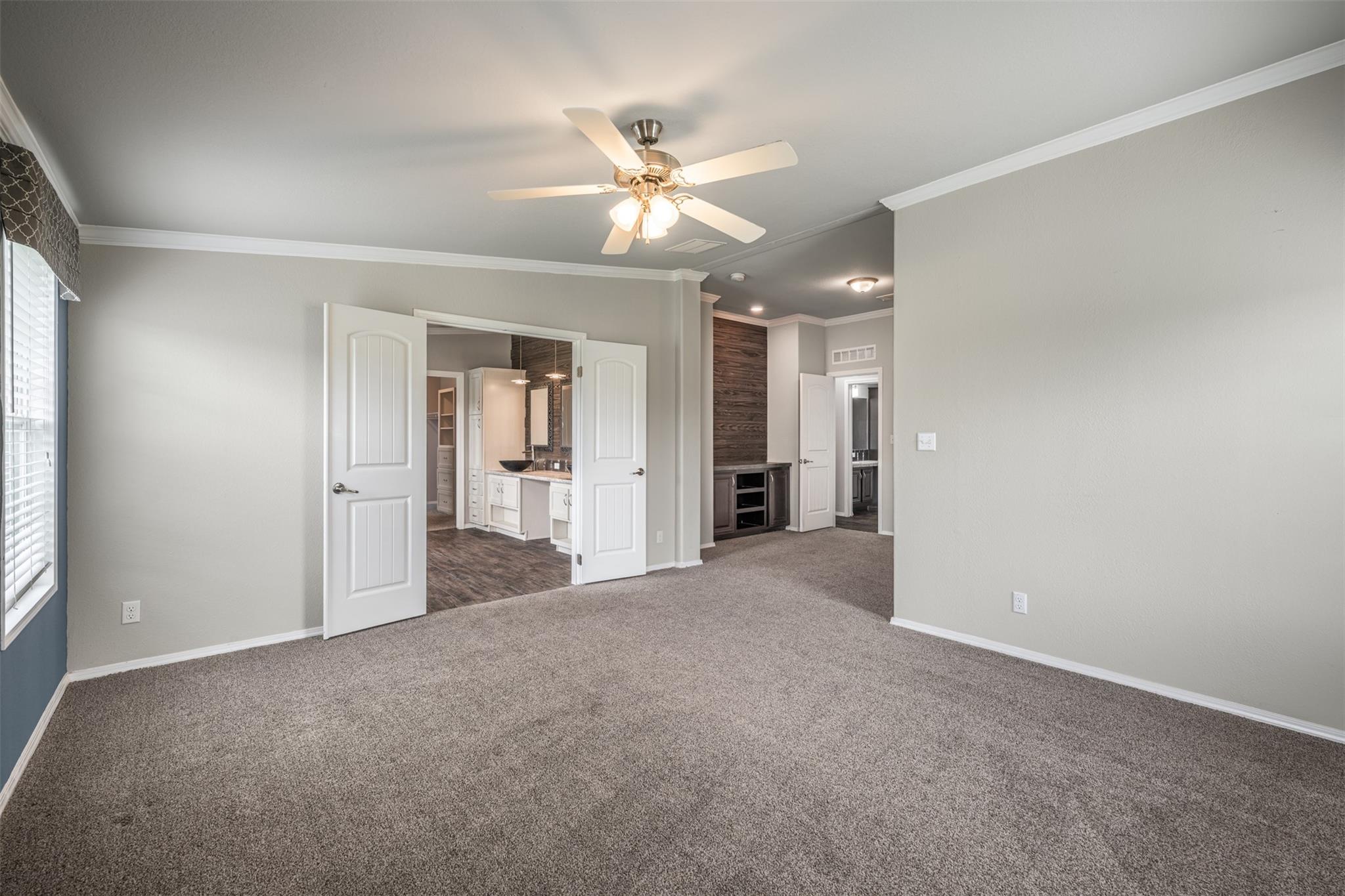 Empty carpeted bedroom with a ceiling fan, open double doors, and adjoining bathroom vanity.