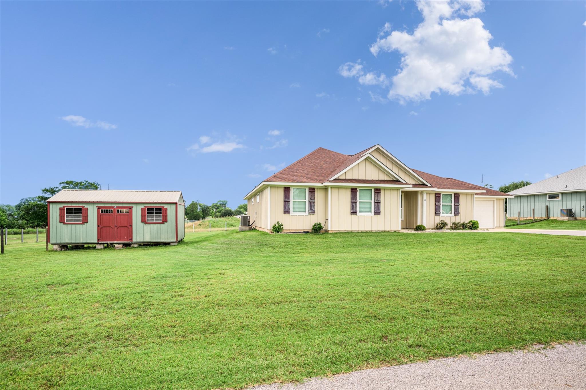 Beige single-story house with a red-roofed green shed beside it on a broad lawn.