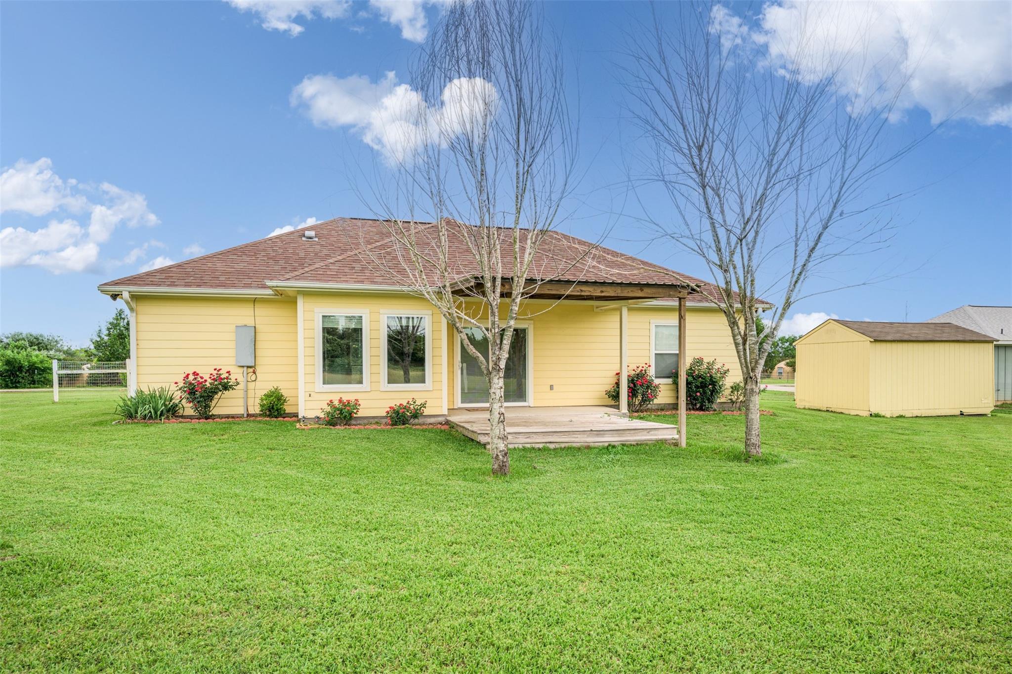 Yellow single-story house with a covered rear deck, leafless trees, green lawn, and matching shed.