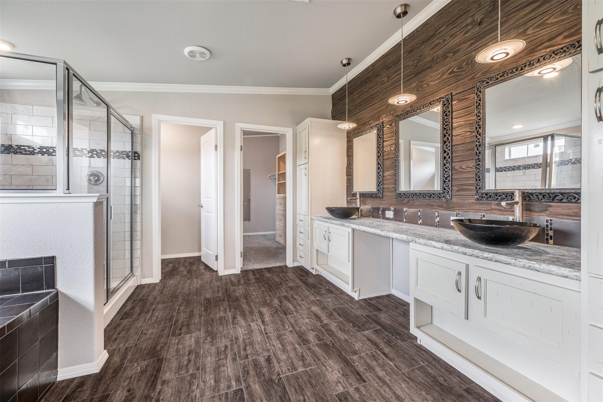 Bathroom with a glass shower and double vanity beneath three mirrors on a wood accent wall.