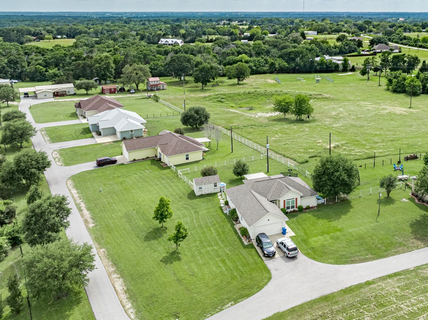 Aerial view of a broad grassy lot and neighboring homes amid wooded countryside.