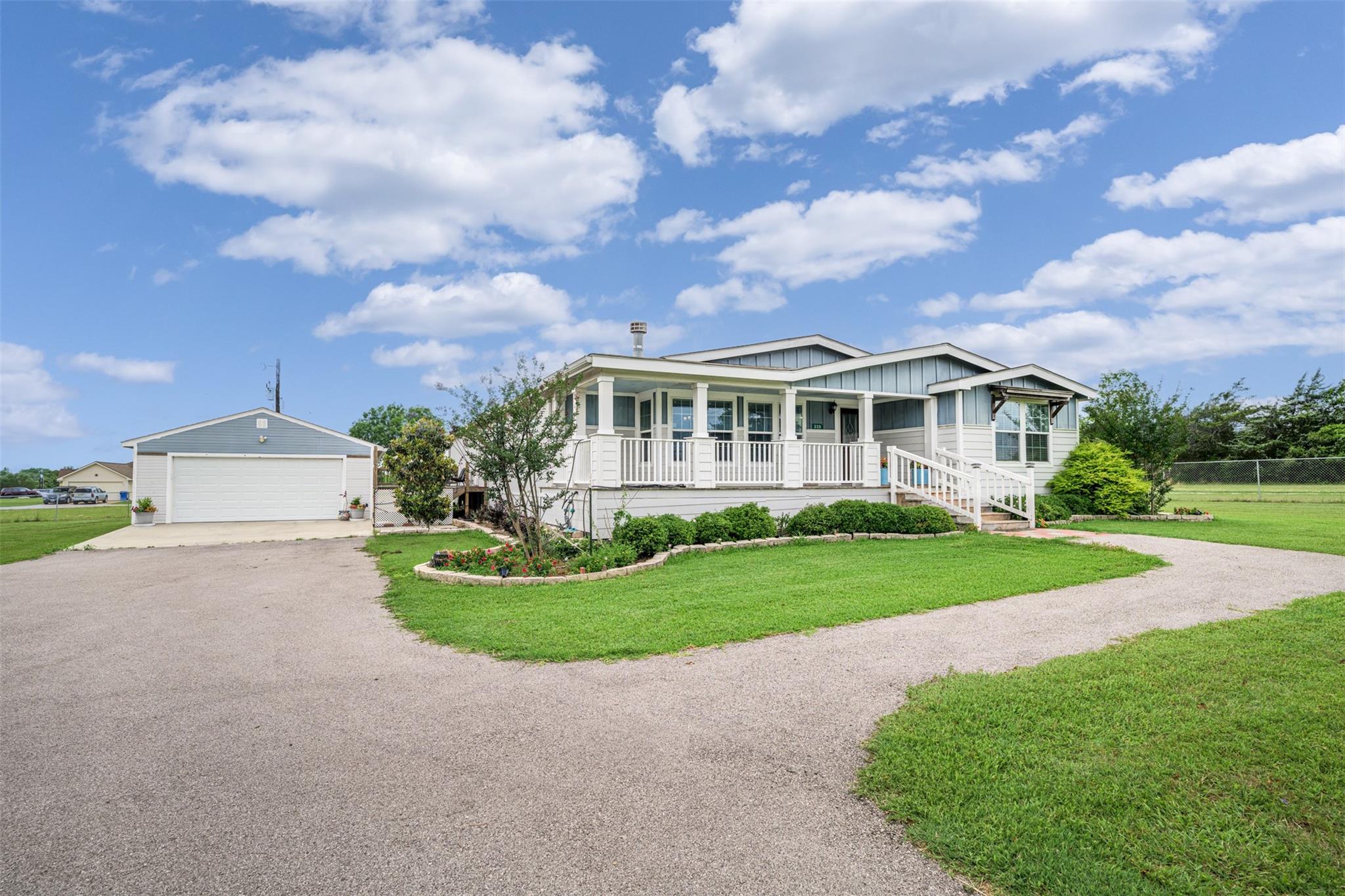 Light-colored home with a raised, white-railed porch beside a detached garage and curving gravel driveway.