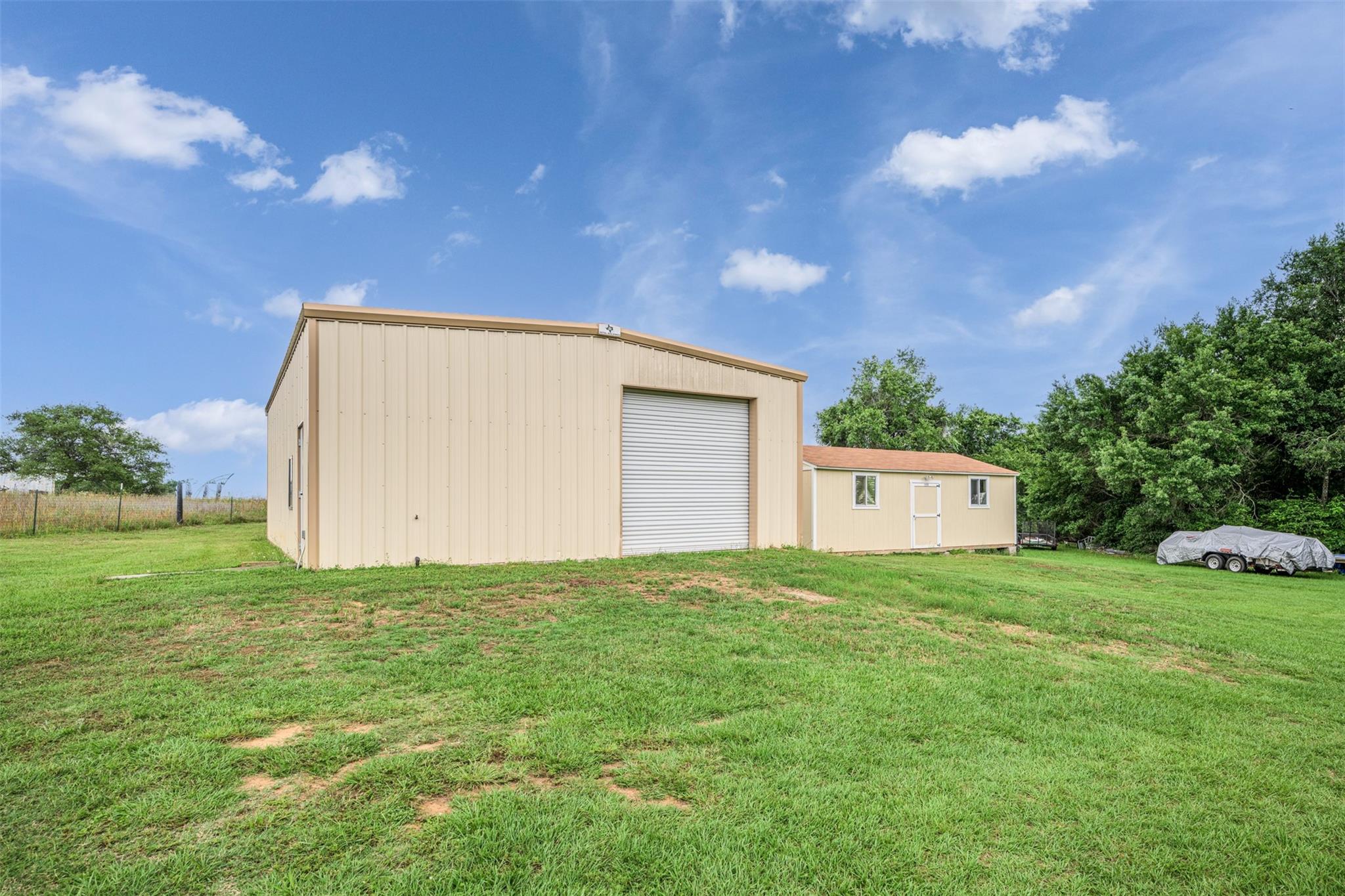 Beige metal workshop with a roll-up door and smaller outbuilding on a grassy property.