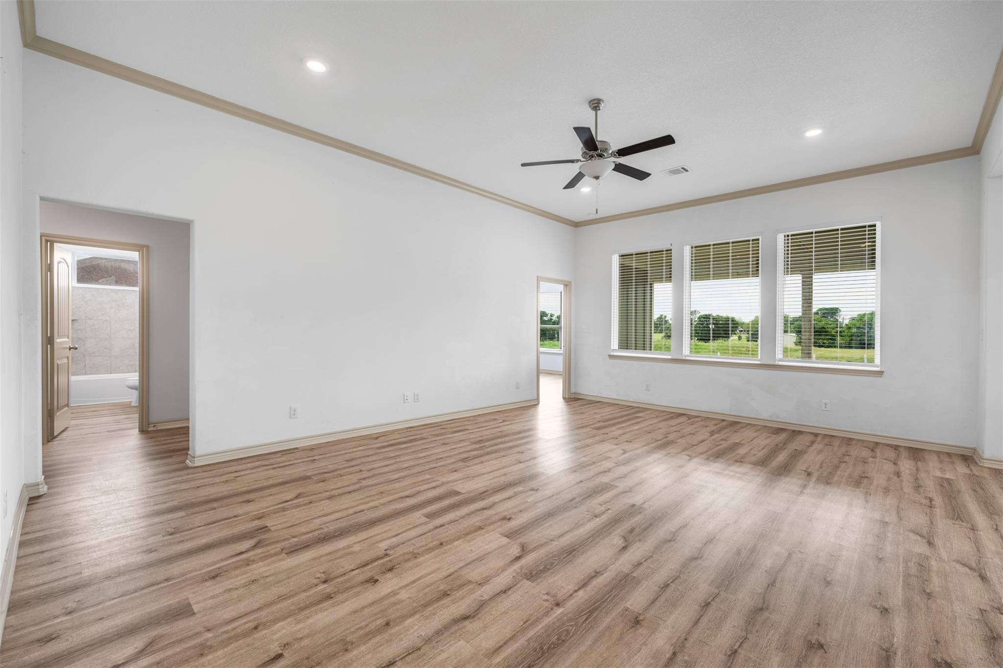 Empty room with wood-look flooring, a tray ceiling fan, three windows, and a bathroom doorway.