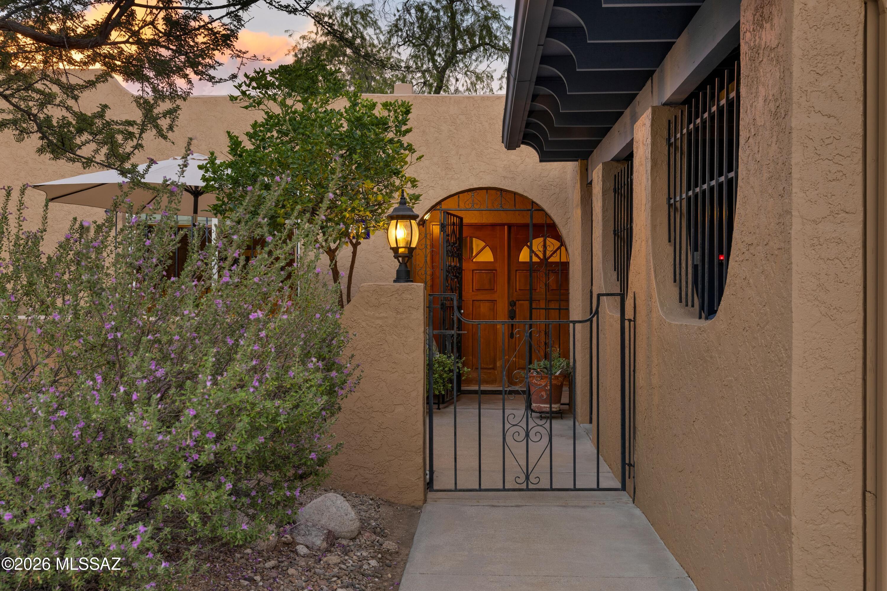 Home entrance with arched wooden doors, wrought-iron gate, and flowering shrubs at dusk.