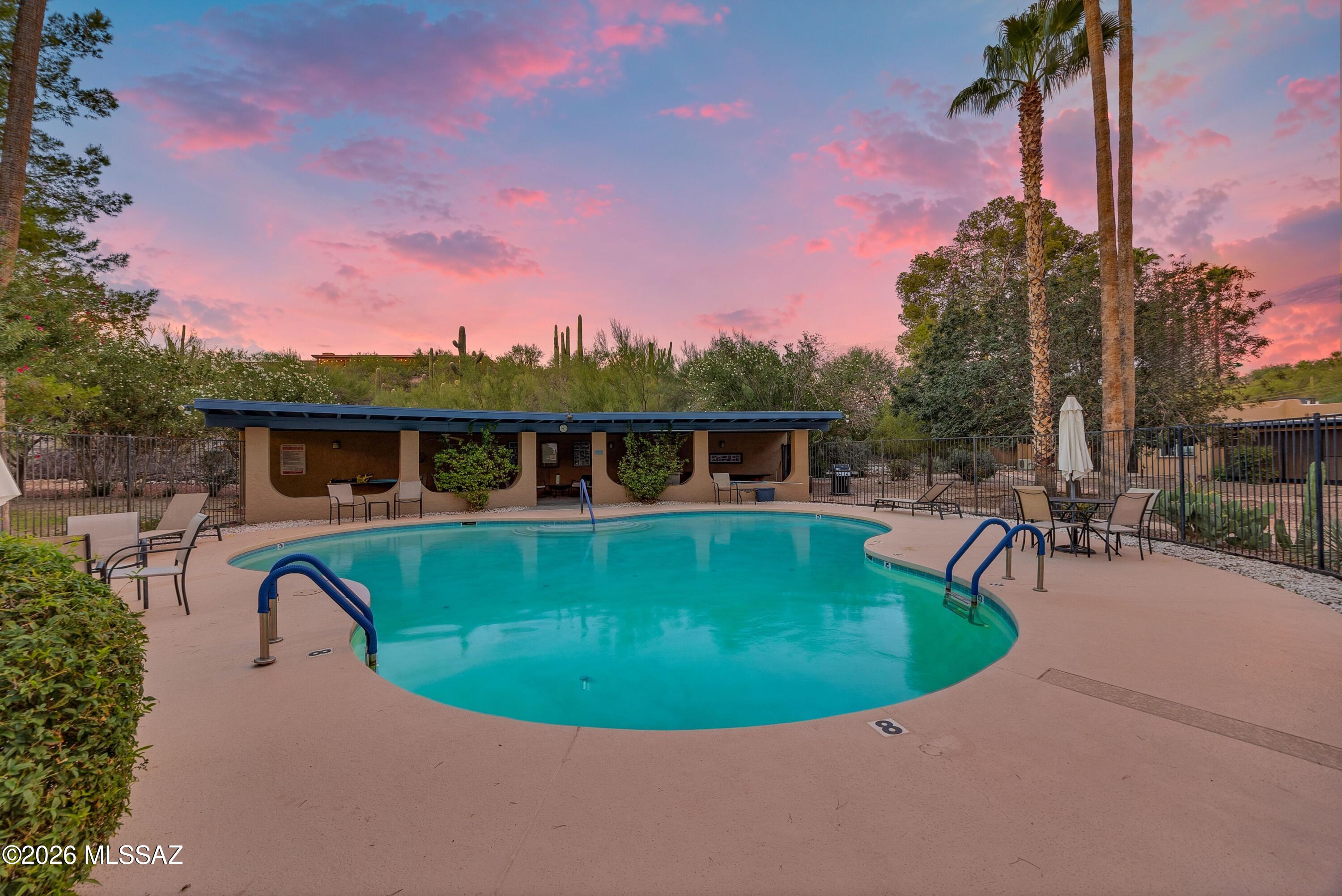 Outdoor pool with curved turquoise water and patio seating beneath a pink sunset sky.