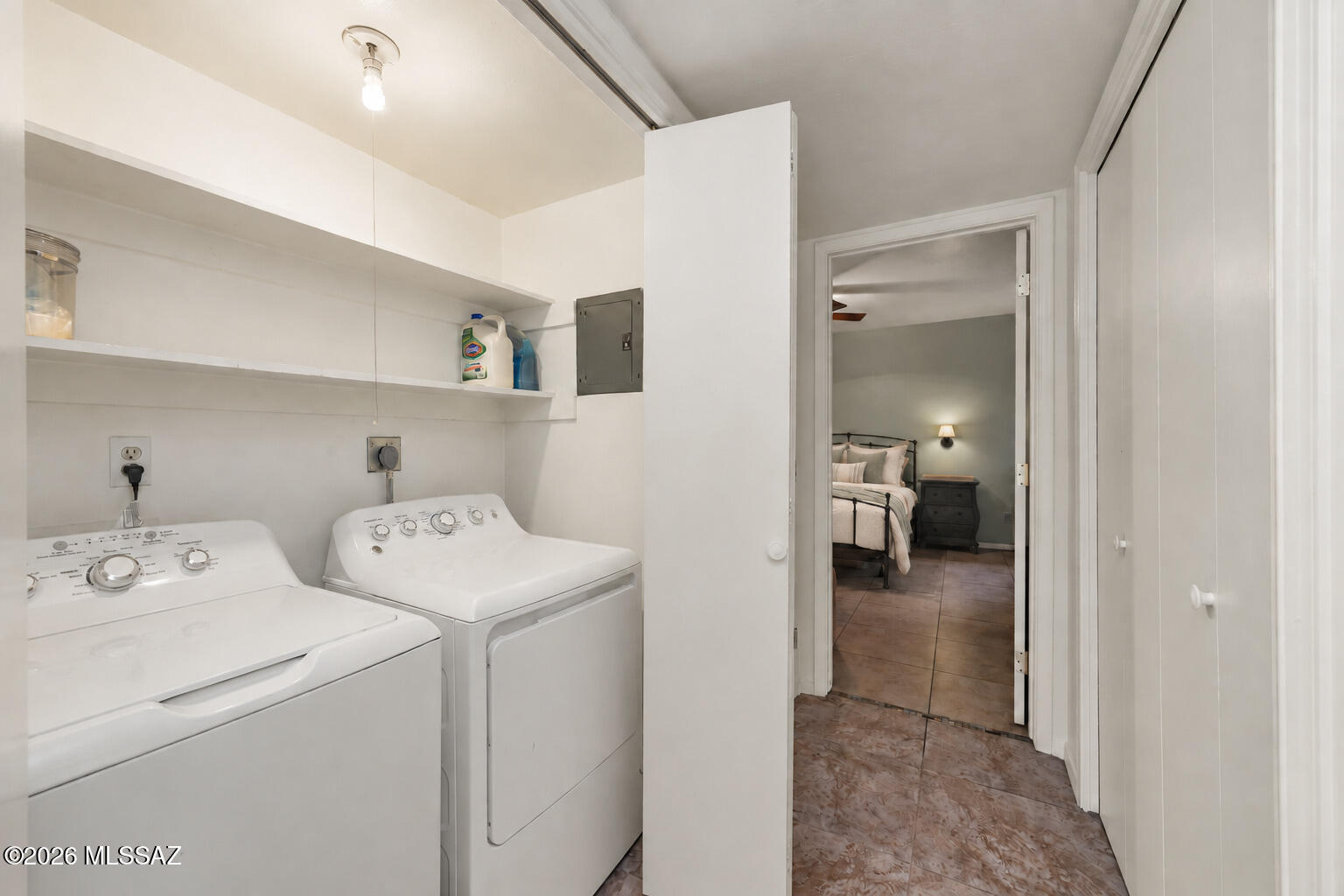 Laundry room with white washer and dryer, open shelves, and a bedroom visible through the doorway.