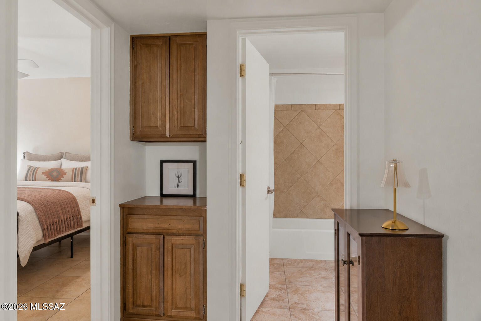 Bathroom with tan tile and tub, beside built-in wooden cabinetry and a bedroom doorway.