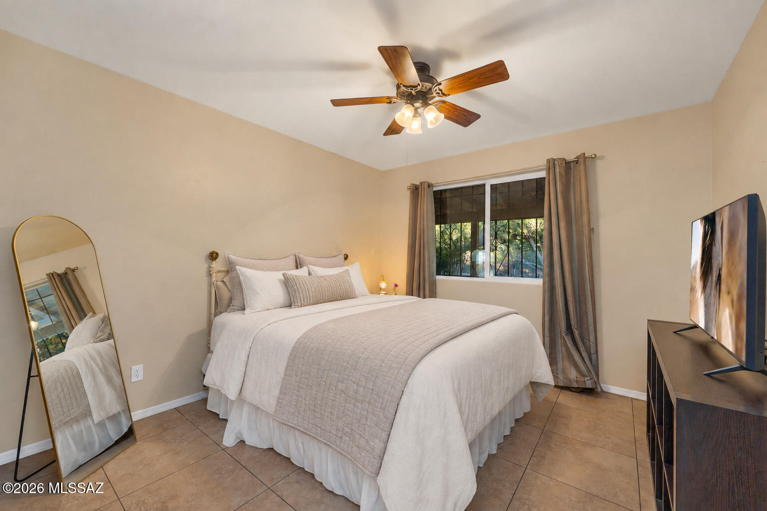Staged bedroom with a white bed, curtained barred window, gold-framed mirror, and ceiling fan.