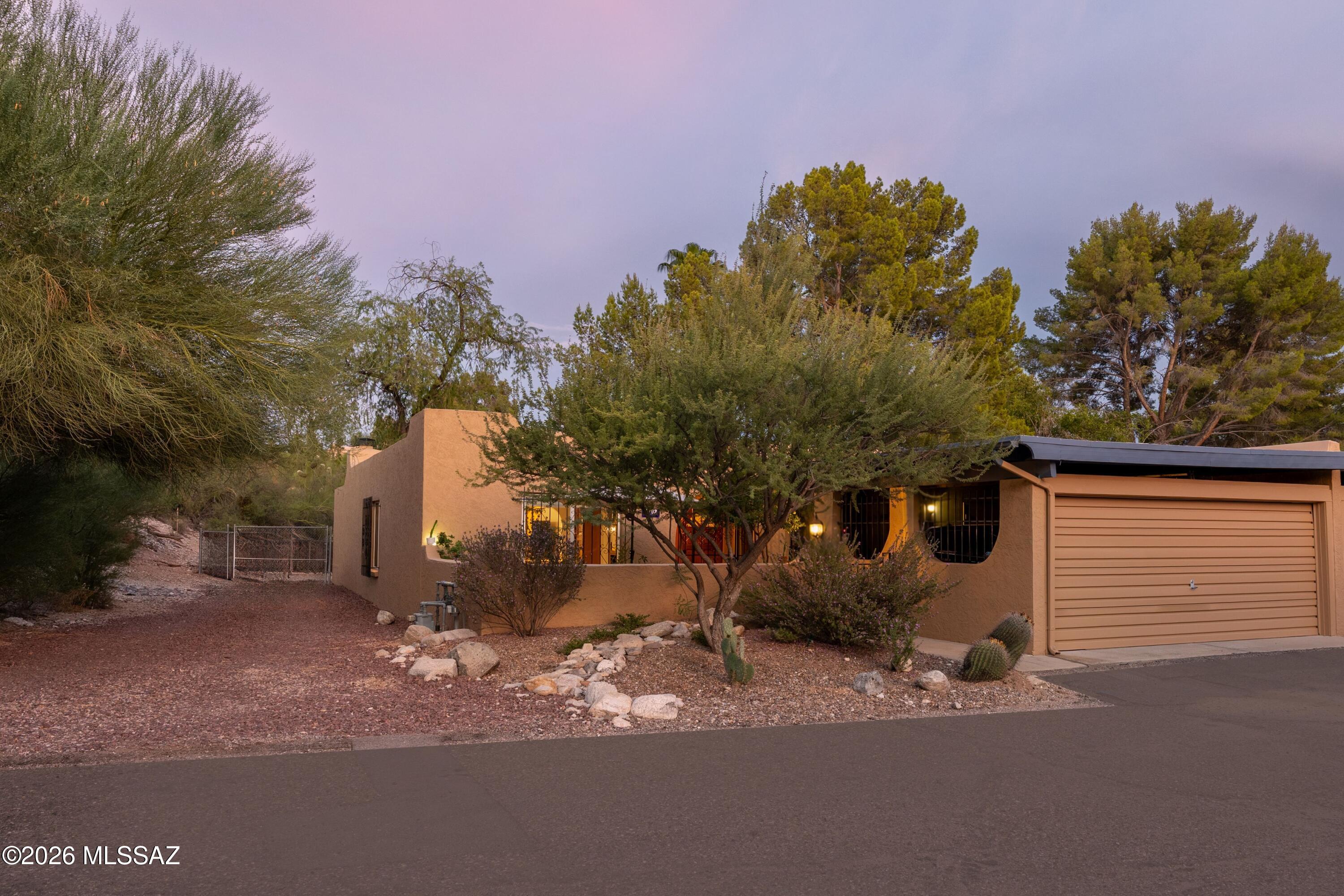 Stucco home with a tan garage, mature trees, and desert landscaping at dusk.