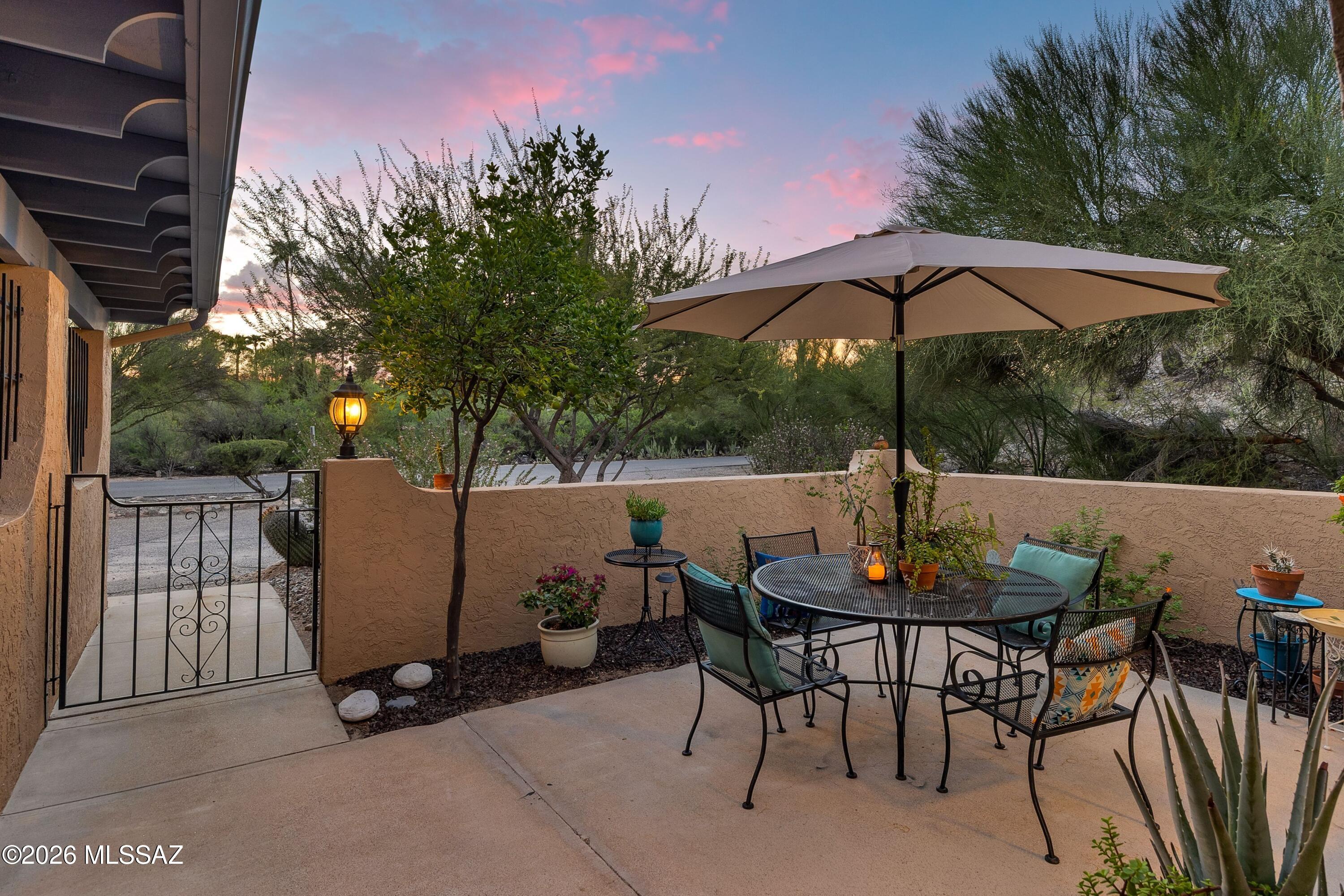 Patio dining area with umbrella, metal table, wrought-iron gate, and desert landscaping at dusk.