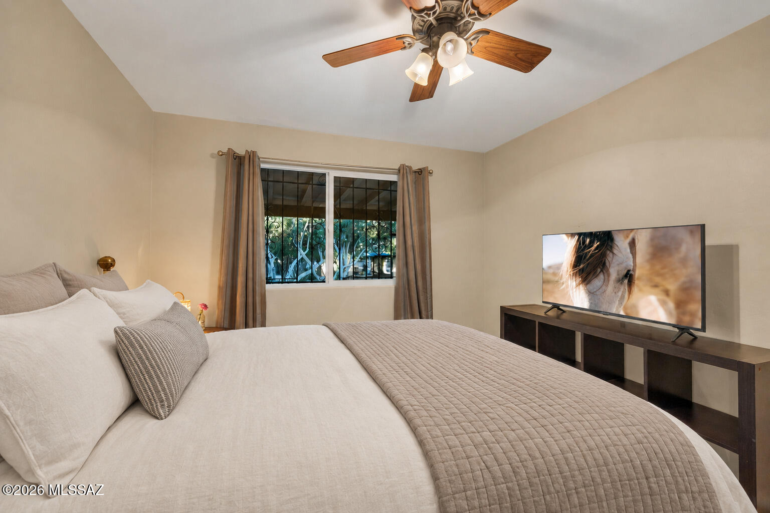 Furnished bedroom with a large white bed, wood-blade ceiling fan, and television.