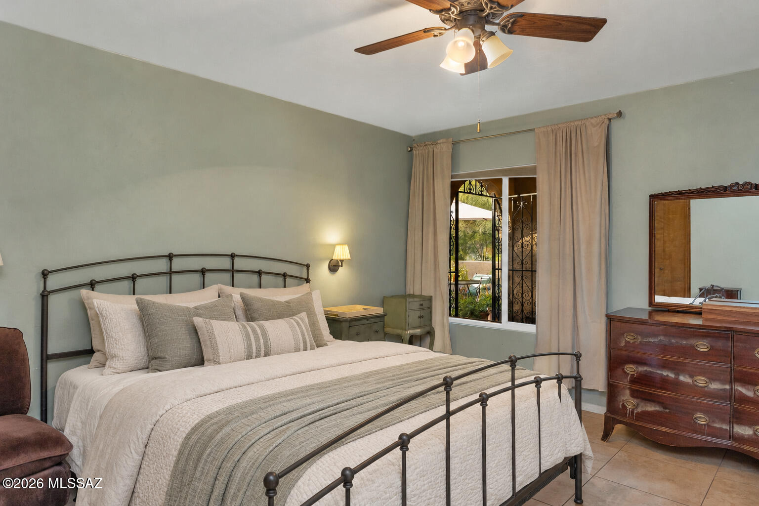 Furnished bedroom with a dark metal bed, ceiling fan, and wooden dresser beside curtained ironwork doors.