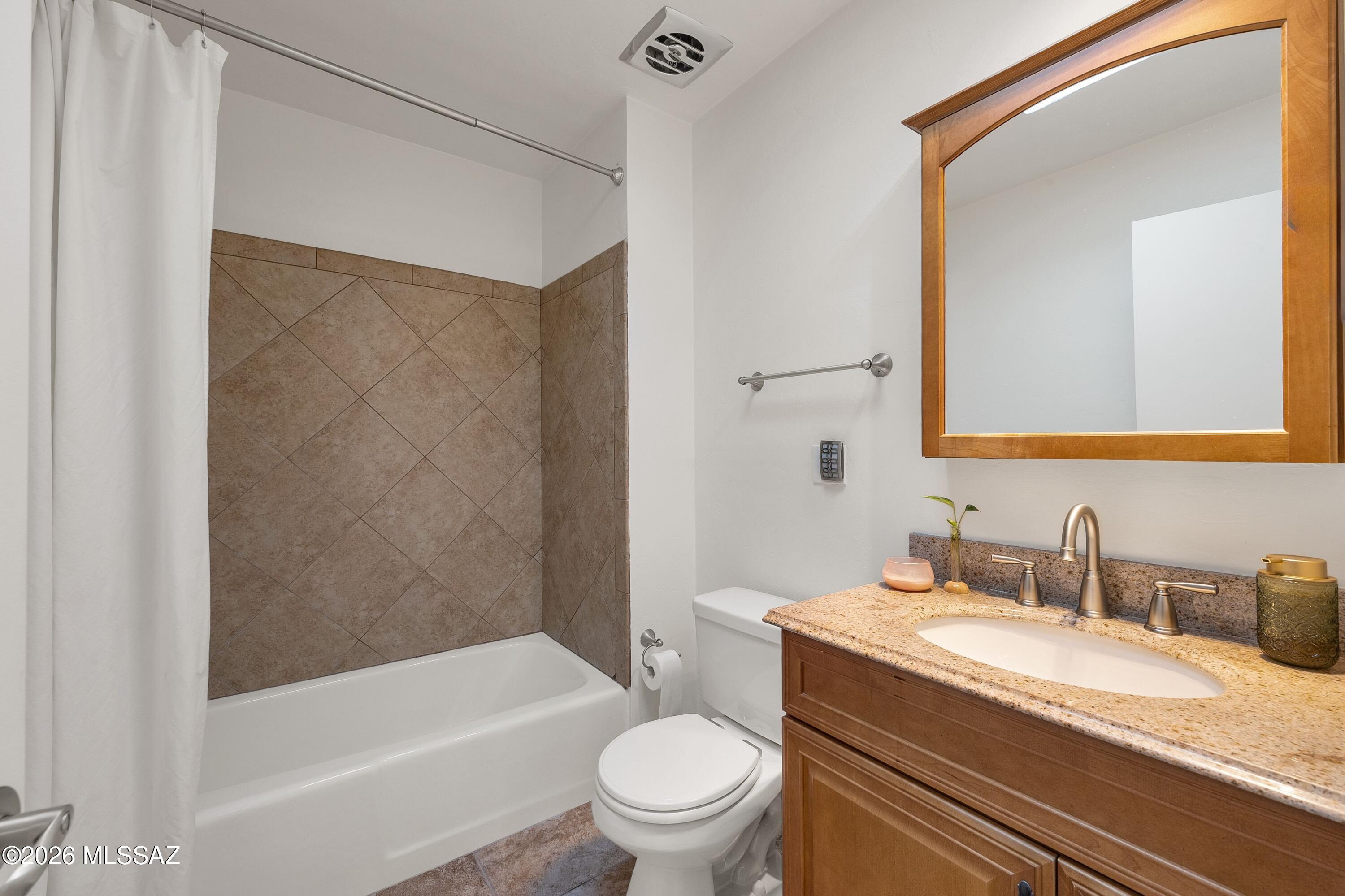 Bathroom with a tiled tub and shower, toilet, and wood vanity topped with granite.