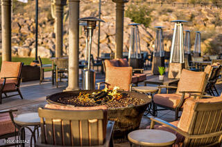 Outdoor patio seating arranged around a fire pit with tall heaters and mature trees.