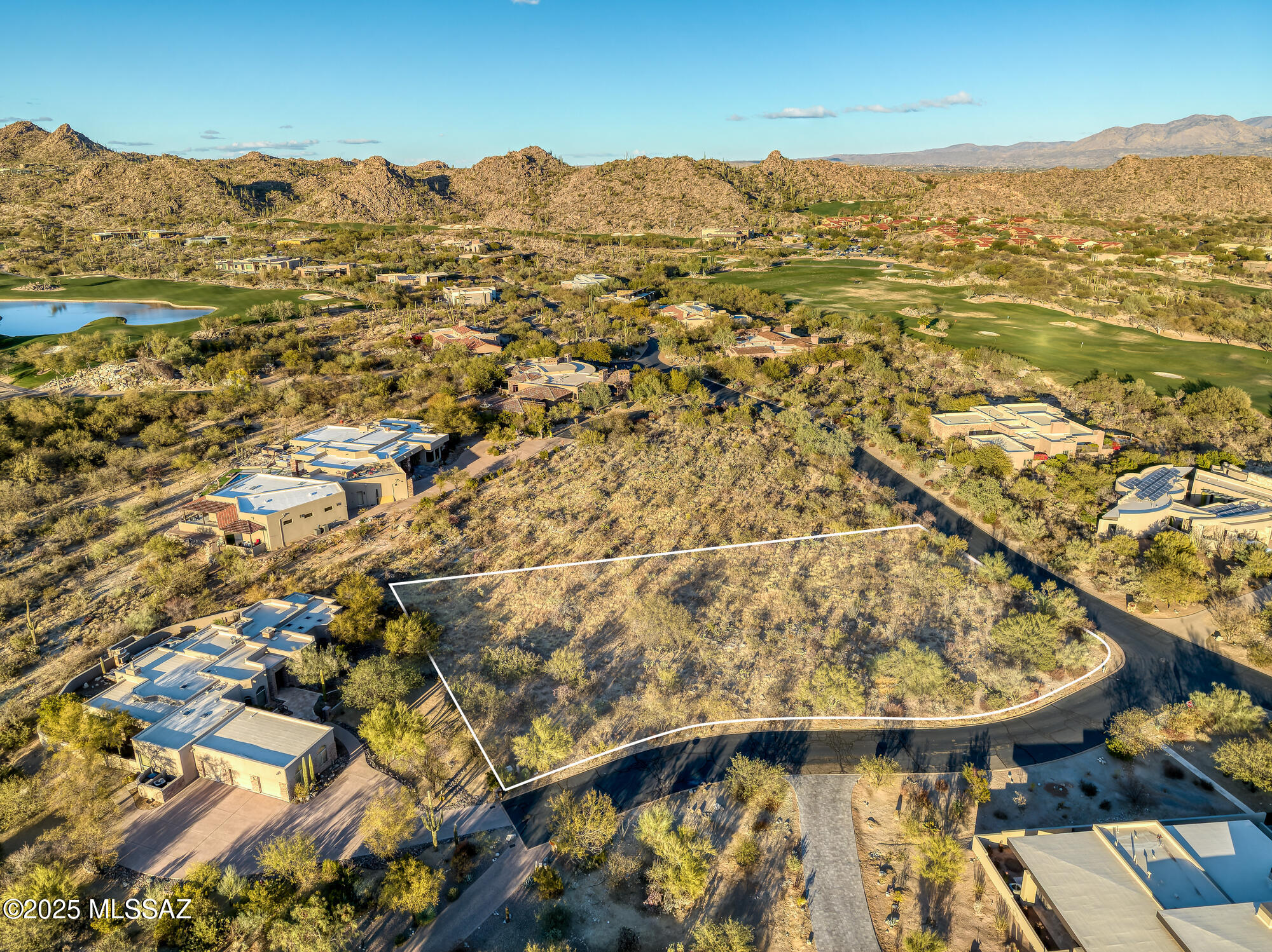 Outlined vacant desert lot beside a golf course, surrounded by homes and distant mountains.