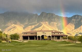 Tan clubhouse beside a green golf course, with rugged mountains and a rainbow behind it.