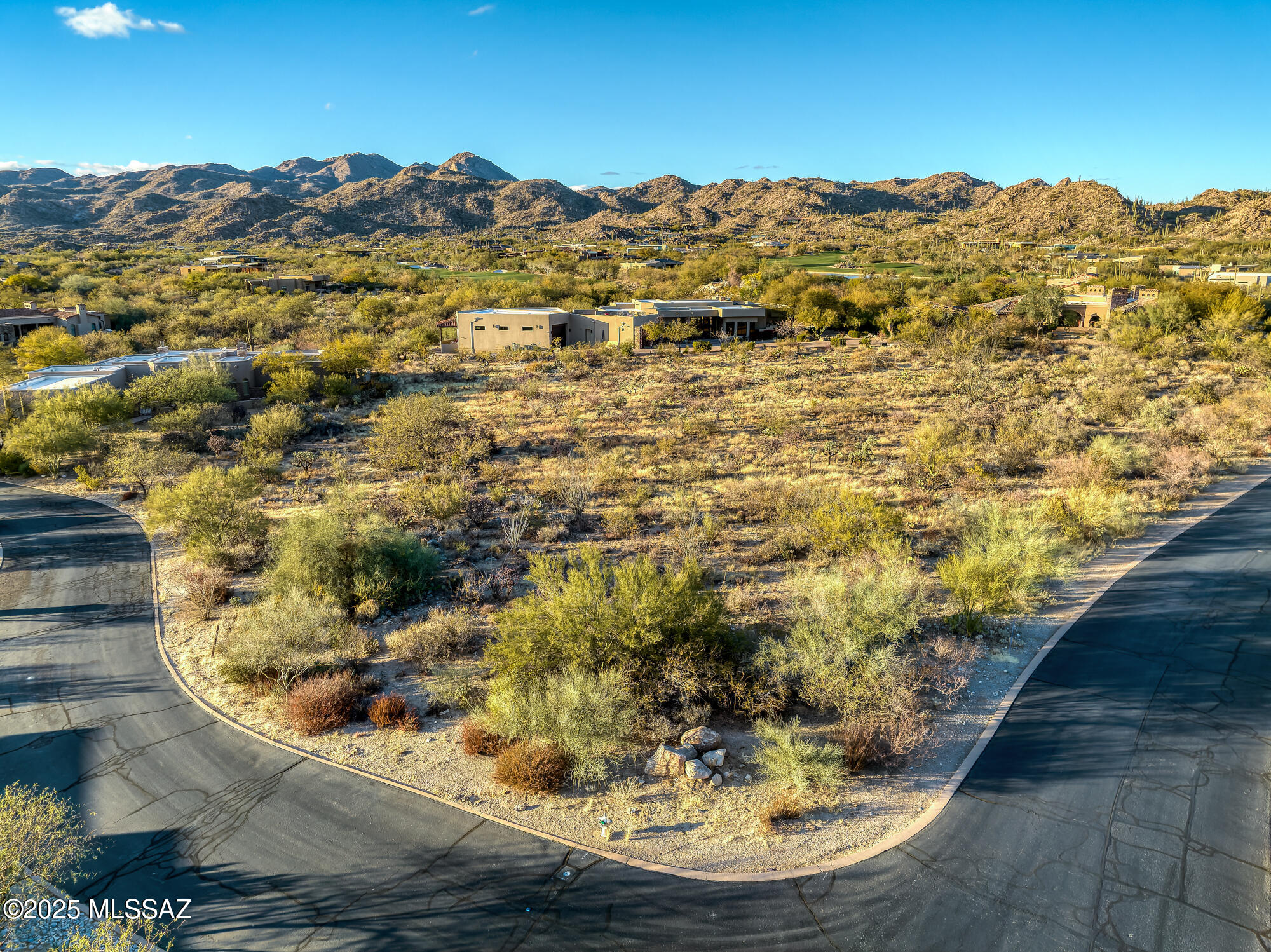 Vacant desert lot at a curved intersection, with native shrubs, nearby homes, and mountain views.