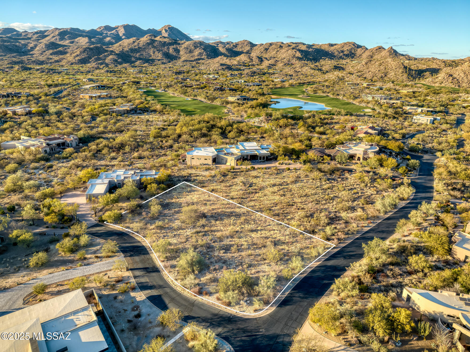 Vacant desert lot outlined in white, with nearby homes, a golf course, pond, and mountains beyond.