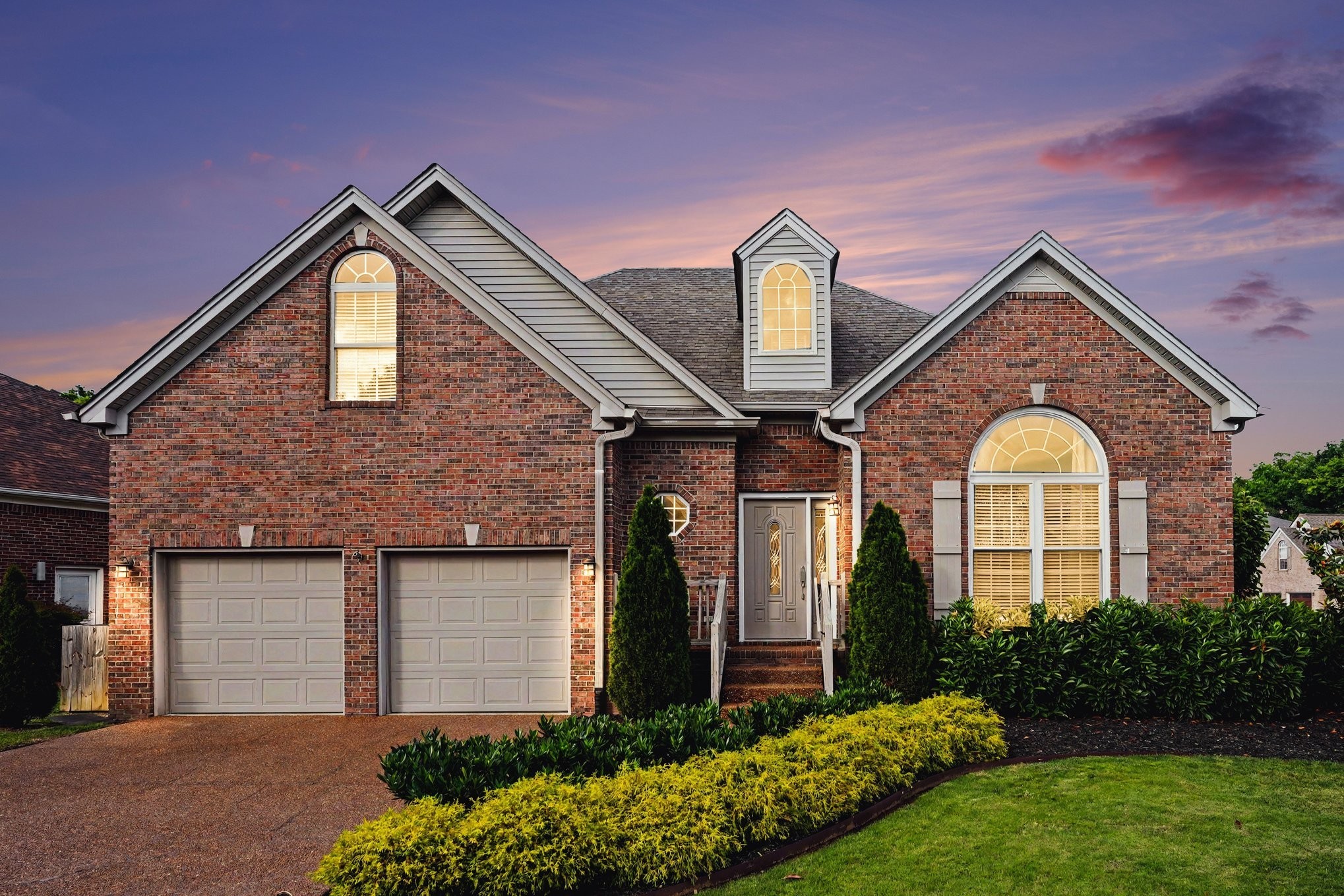 Red brick home with two garages, arched windows, and landscaped front yard at dusk.