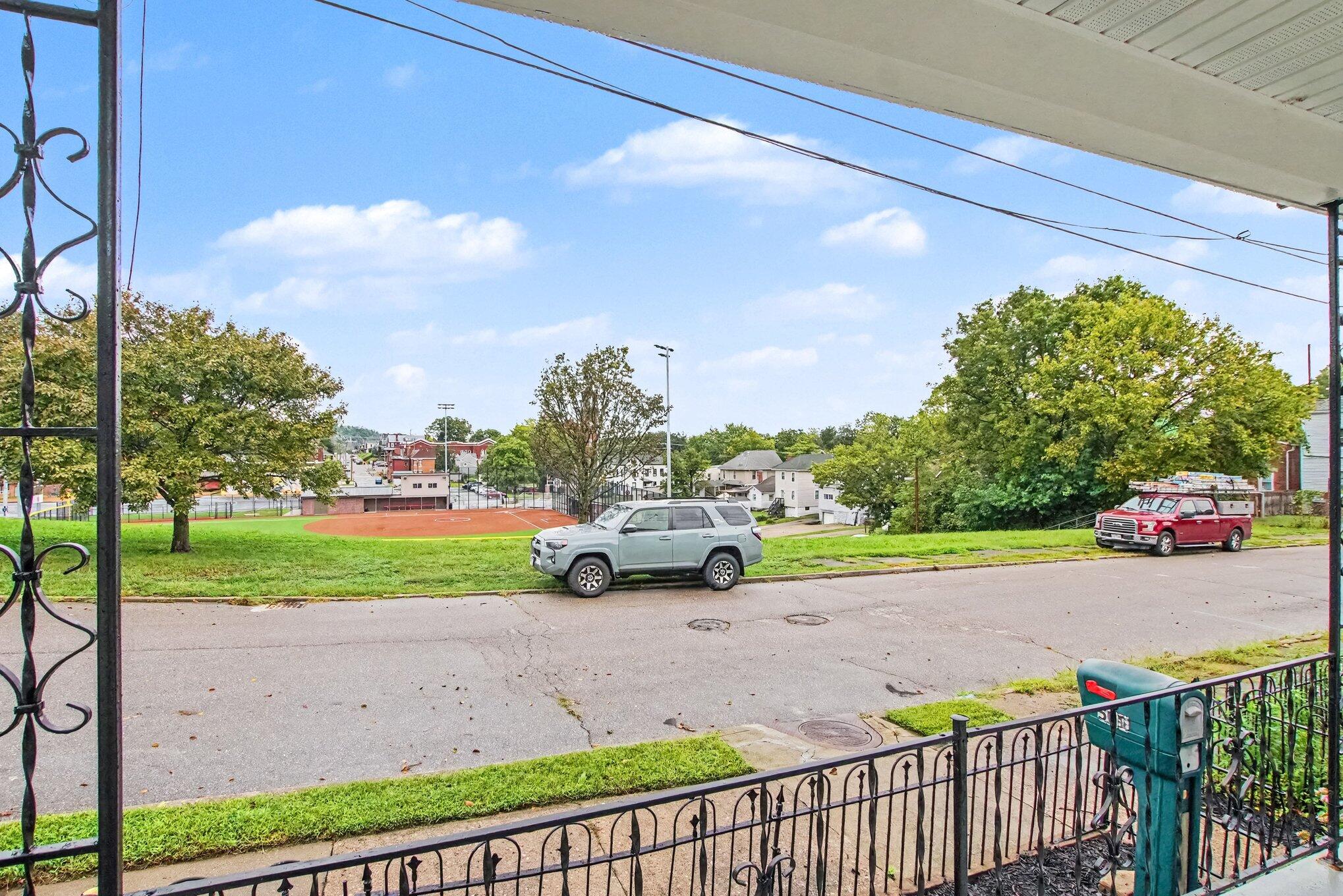 Porch view across the street to a neighborhood baseball field, trees, and nearby homes.