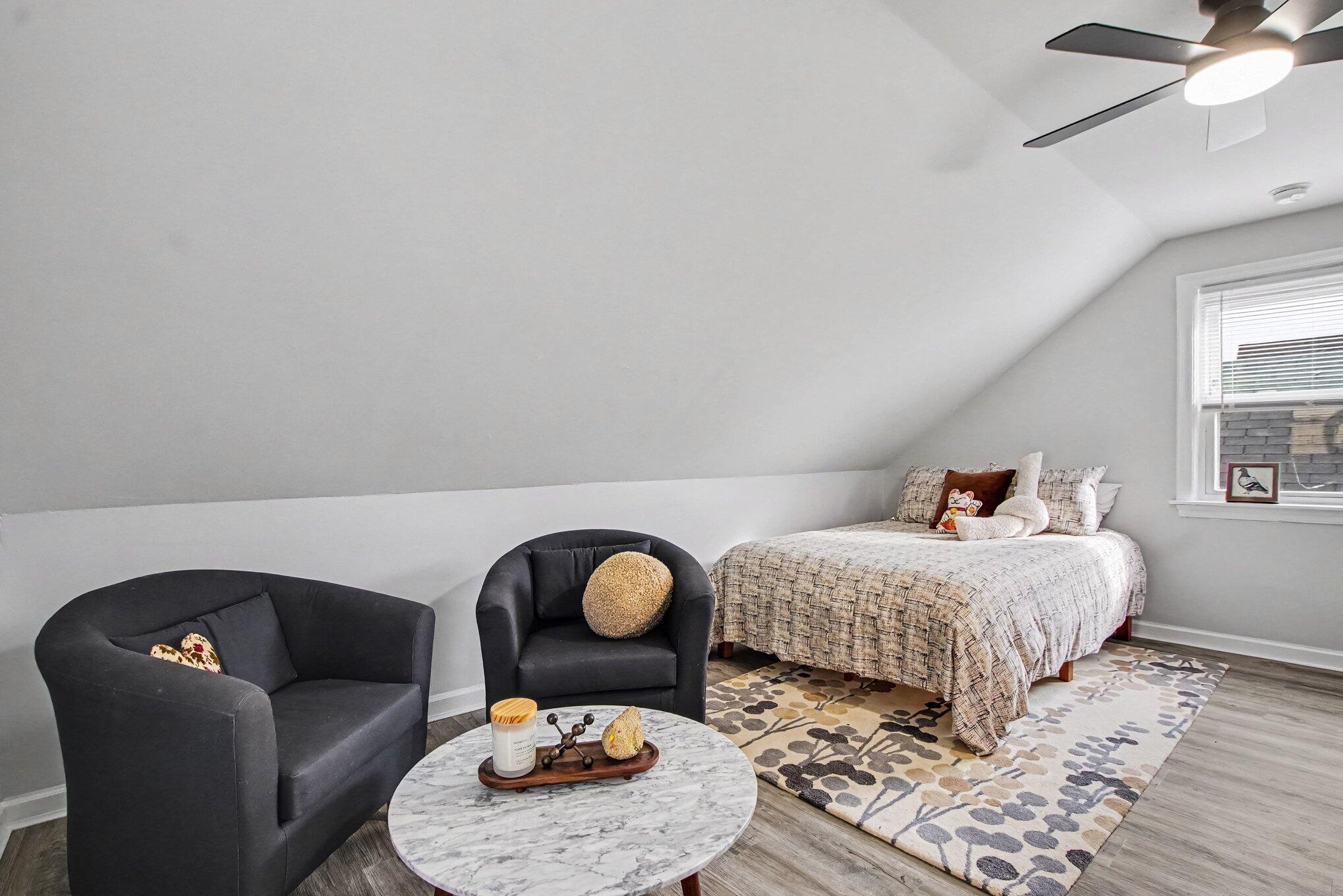 Furnished attic bedroom with a sloped ceiling, patterned bedspread, dark chairs, and round marble coffee table.