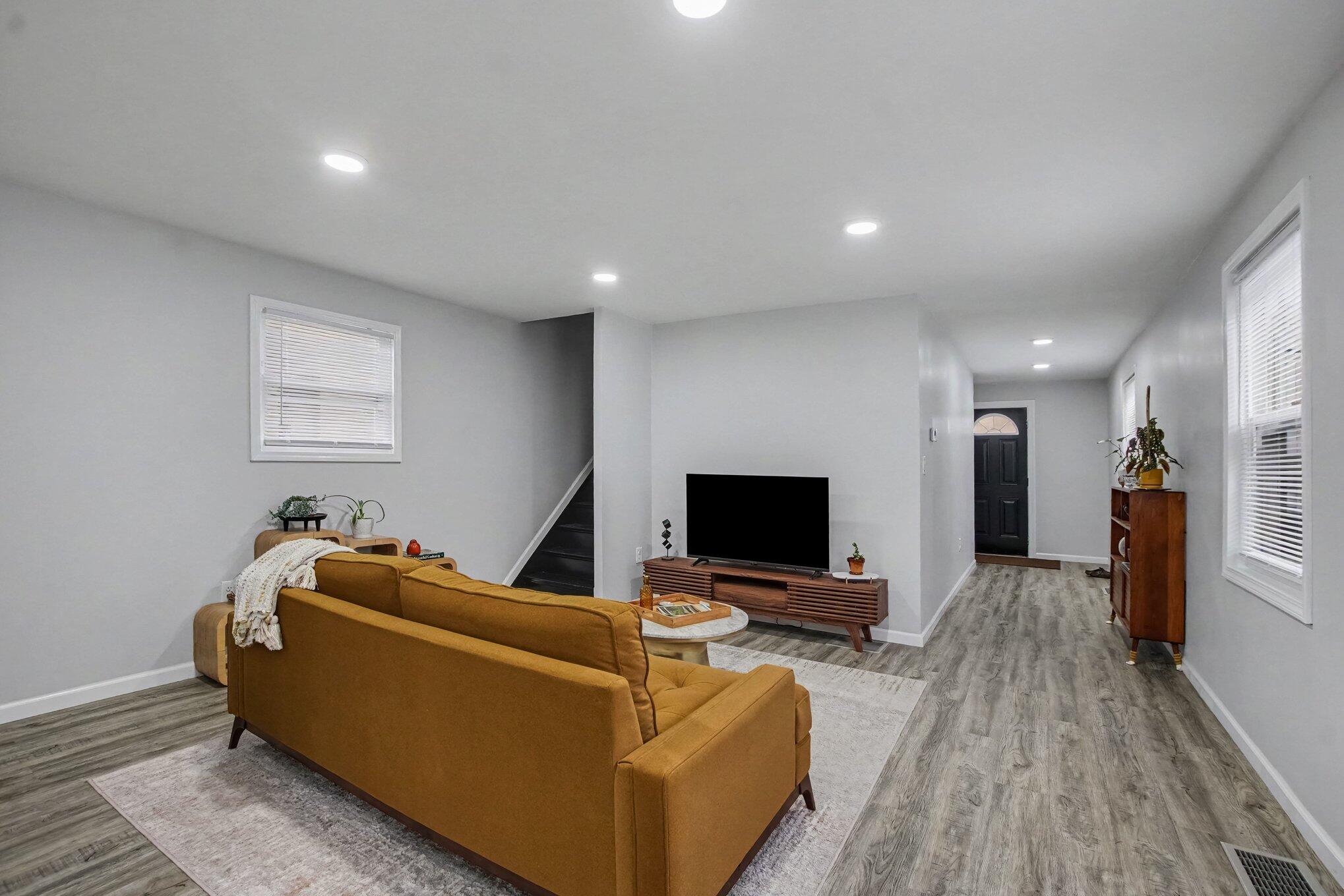 Living room with a mustard sectional, television, gray plank flooring, and a hallway to the front door.