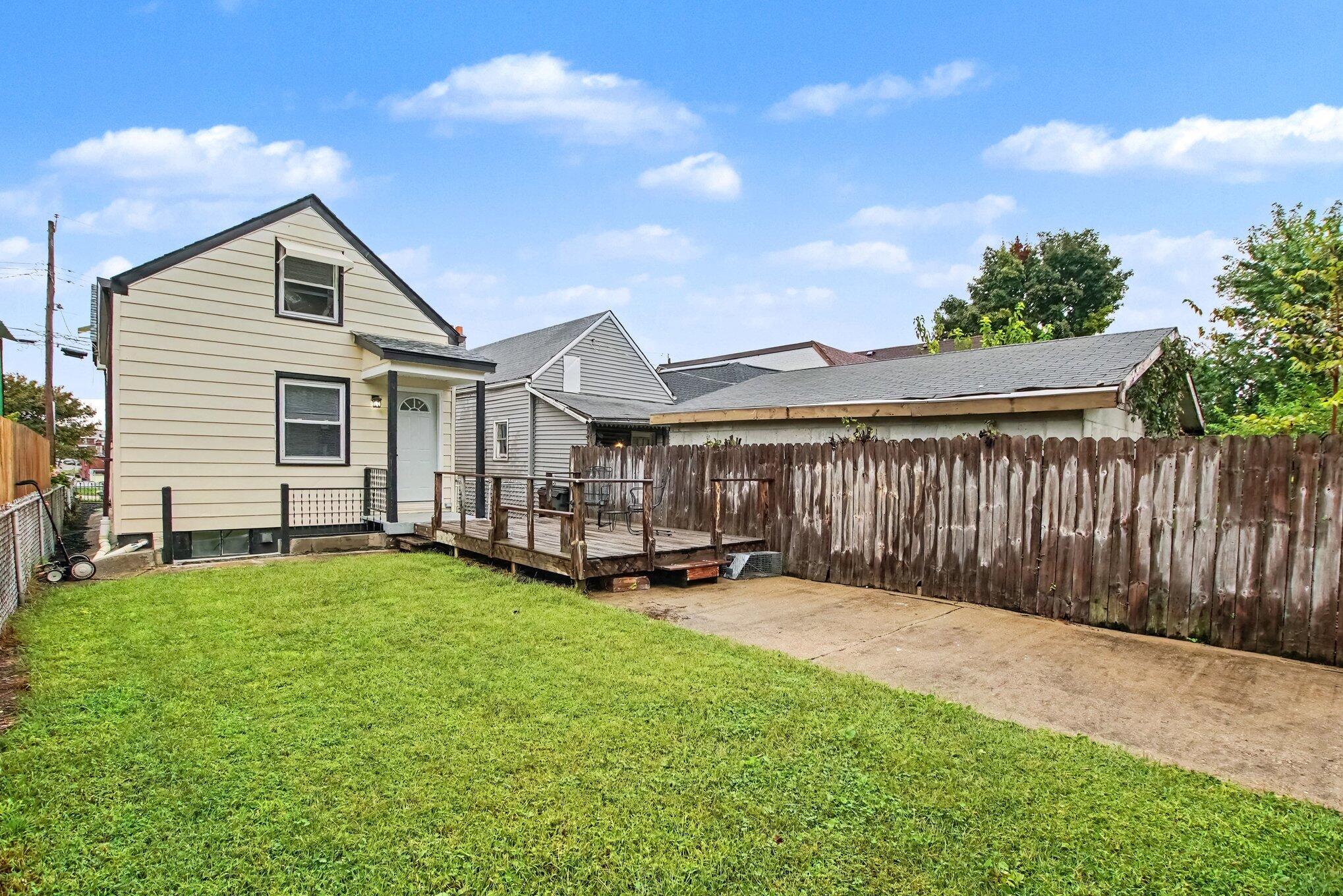 Rear exterior of a cream-sided house with a small deck, fenced lawn, and concrete driveway.
