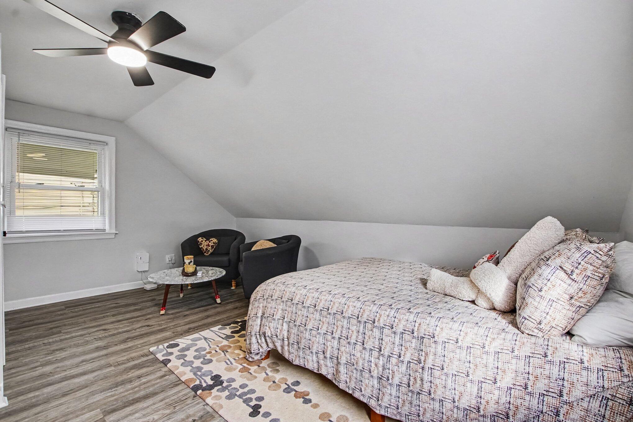 Furnished bedroom with a sloped ceiling, patterned bed, ceiling fan, and small seating area.