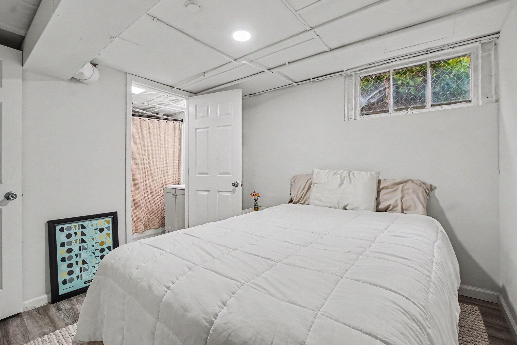 Basement bedroom with a white bed, small high window, and open doorway to a curtained area.