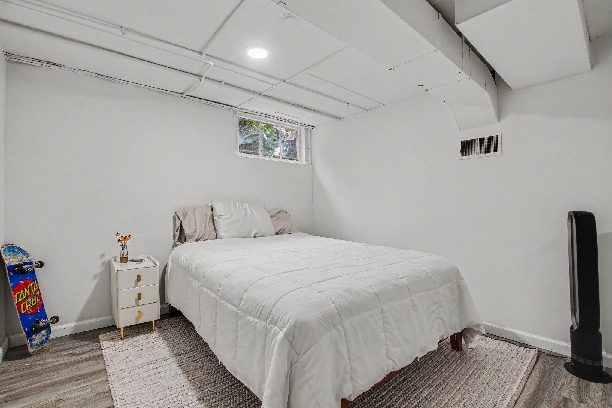 Bedroom with a white bed, small high window, and skateboard leaning against the wall.