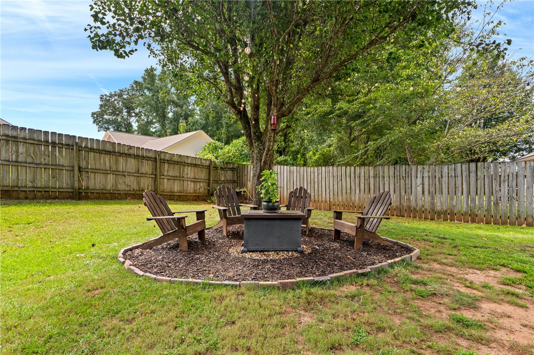 Fenced backyard seating area with Adirondack chairs around a fire pit beneath a large tree.