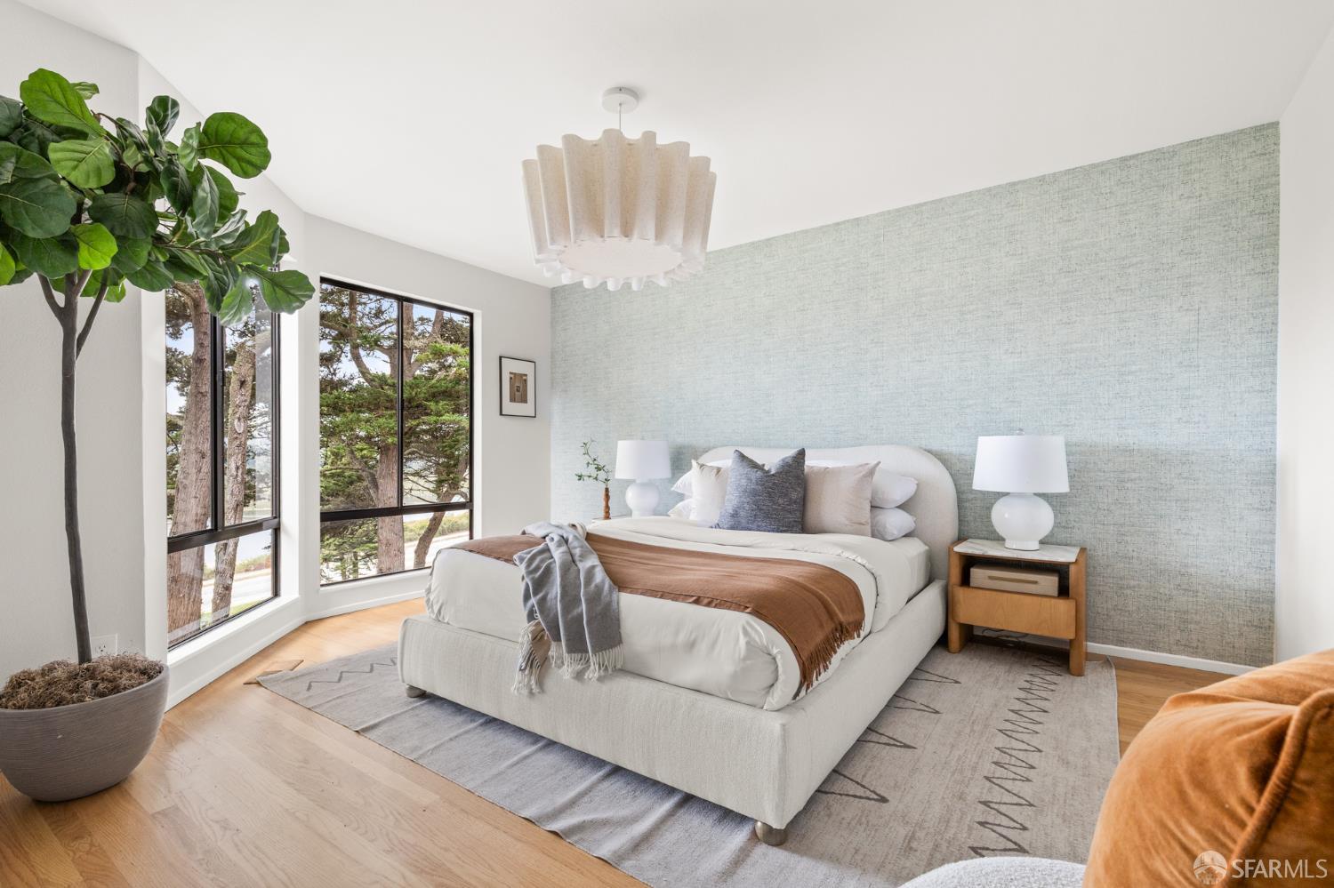 Staged bedroom with a white bed, pale blue accent wall, and large windows overlooking trees.