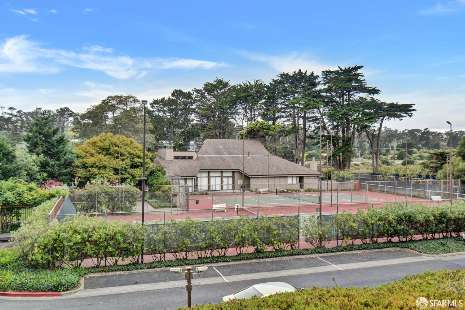 Outdoor tennis courts beside a low clubhouse, framed by mature trees and landscaped grounds.