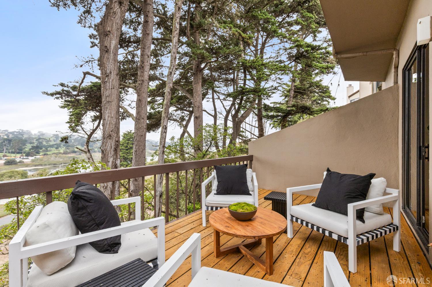Furnished wooden balcony overlooking trees and water, with white lounge chairs and a round coffee table.