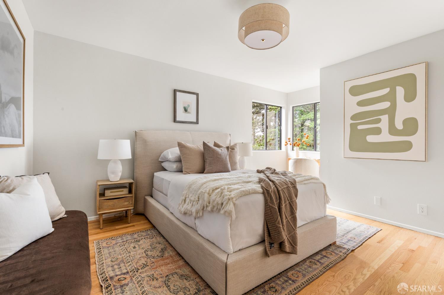 Staged bedroom with a neutral upholstered bed, patterned rugs, hardwood floors, and windows overlooking greenery.