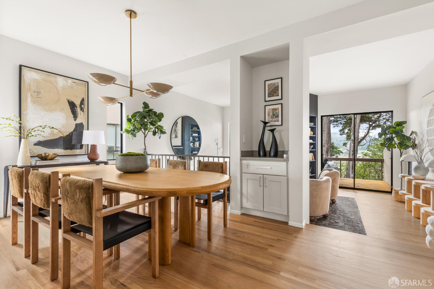 Dining room with a large oval wood table, upholstered chairs, and glass doors opening to a tree-lined balcony.