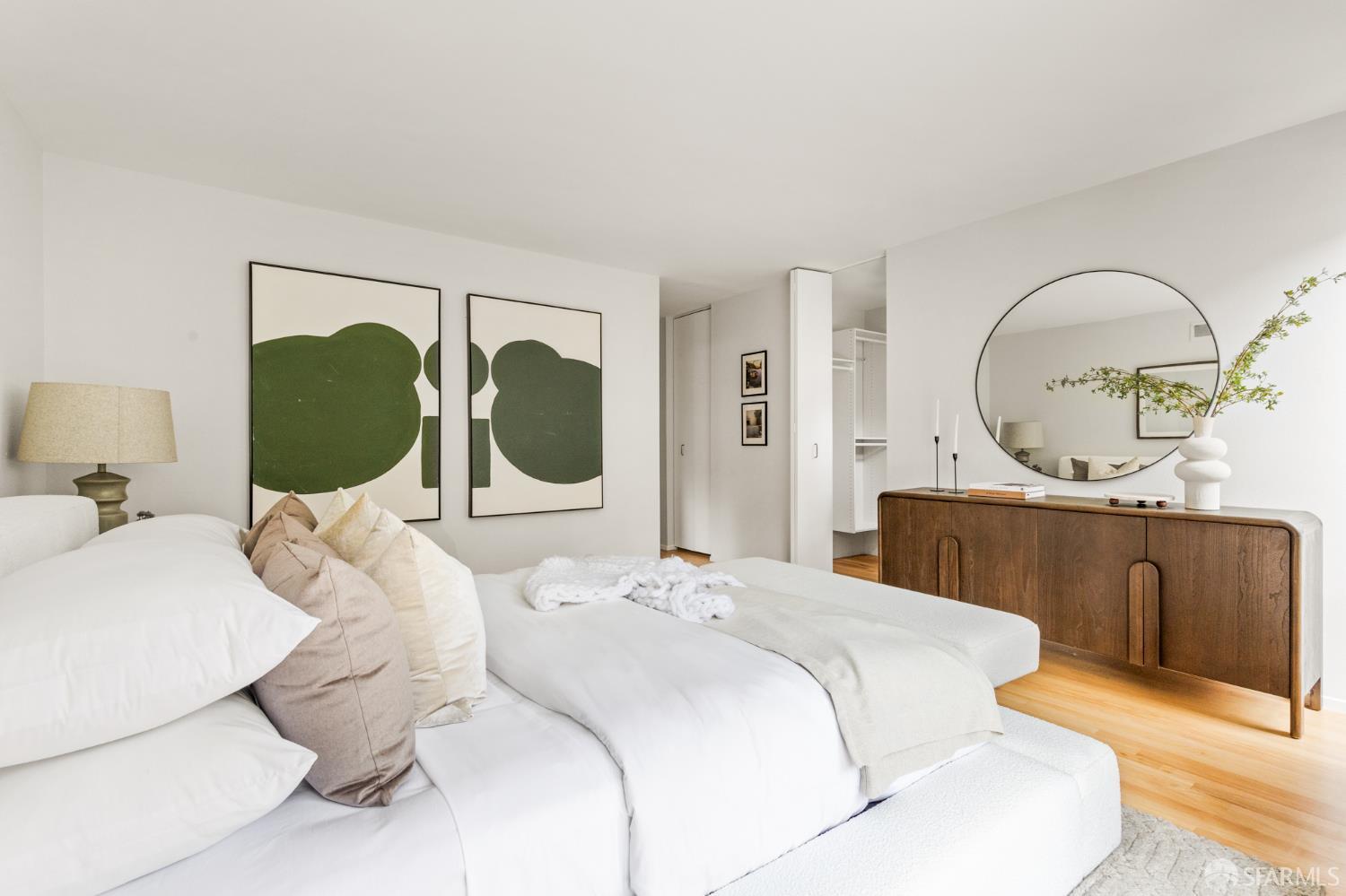 Staged bedroom with a white bed, green abstract artwork, round mirror, and wood dresser.