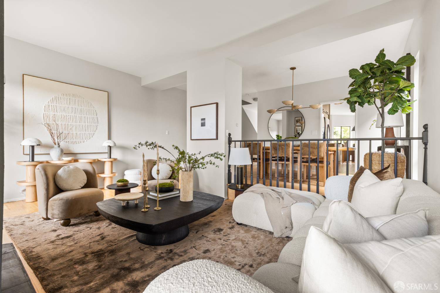 Staged living room with a white sectional, dark sculptural coffee table, and dining area beyond a black railing.