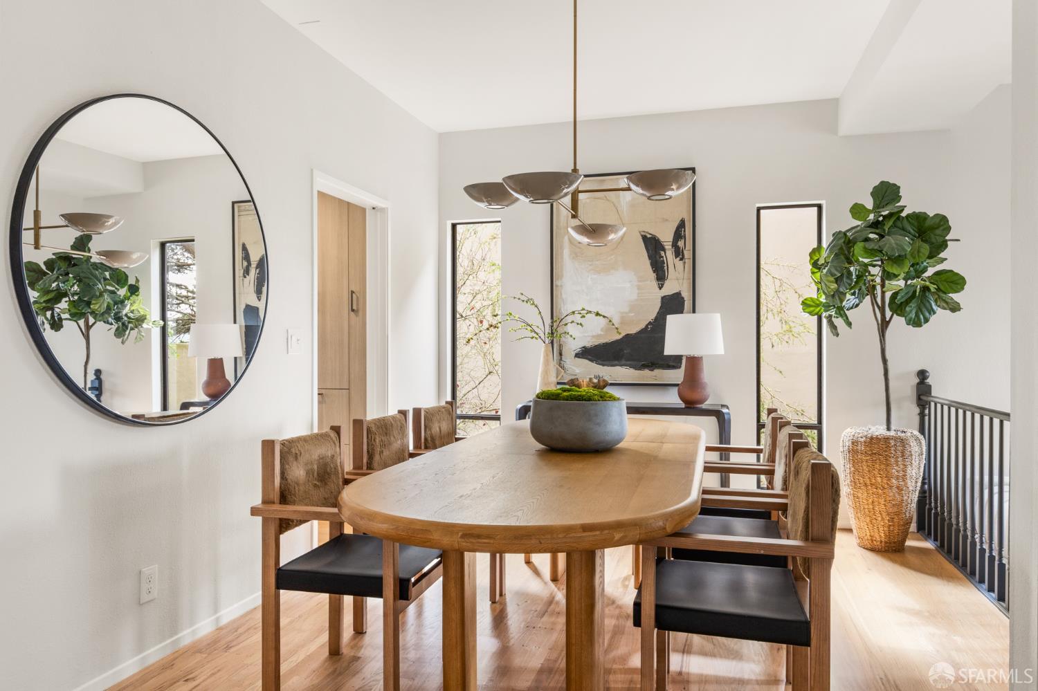 Dining room with a wooden table, upholstered chairs, large round mirror, and tall windows.