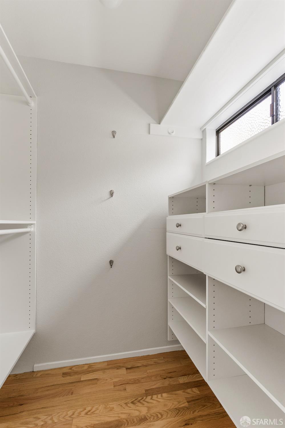 Empty walk-in closet with white built-in drawers and shelves, high window, and hardwood floors.