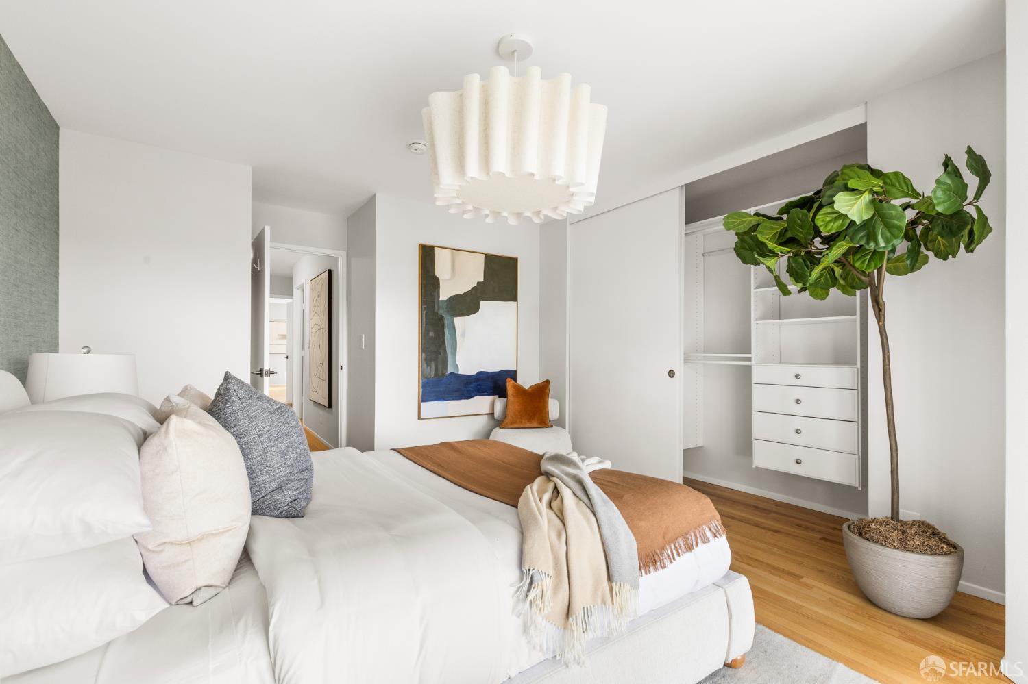 Staged bedroom with a layered white bed, open built-in closet, and large potted tree.