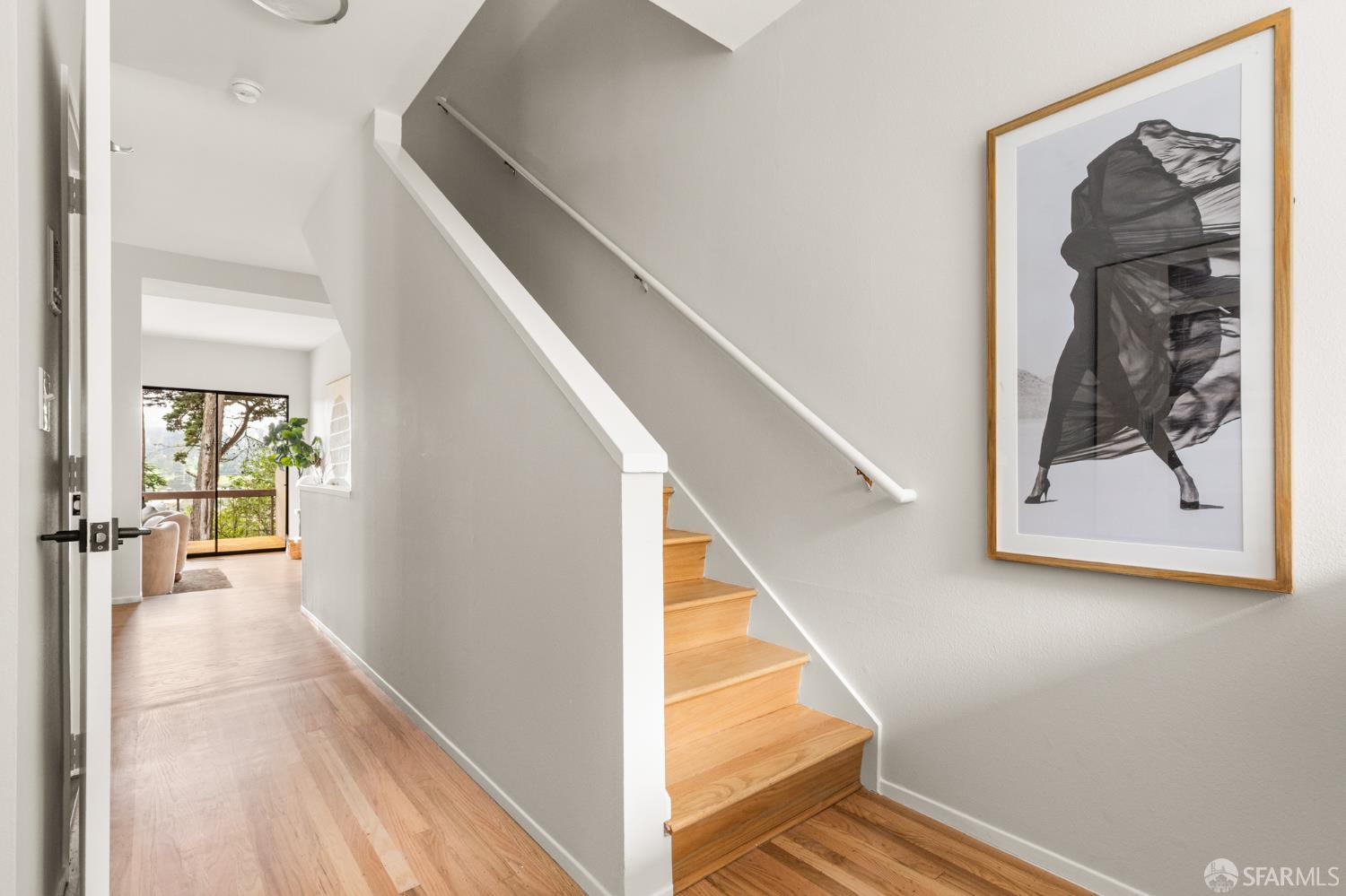 Hallway with a wood staircase, framed black-and-white artwork, and bright sitting area beyond.