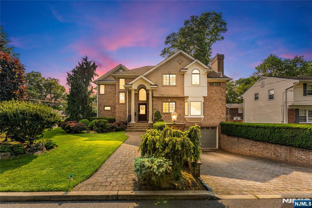 Two-story brick and stucco house with illuminated windows, landscaped lawn, and paved driveway at dusk.